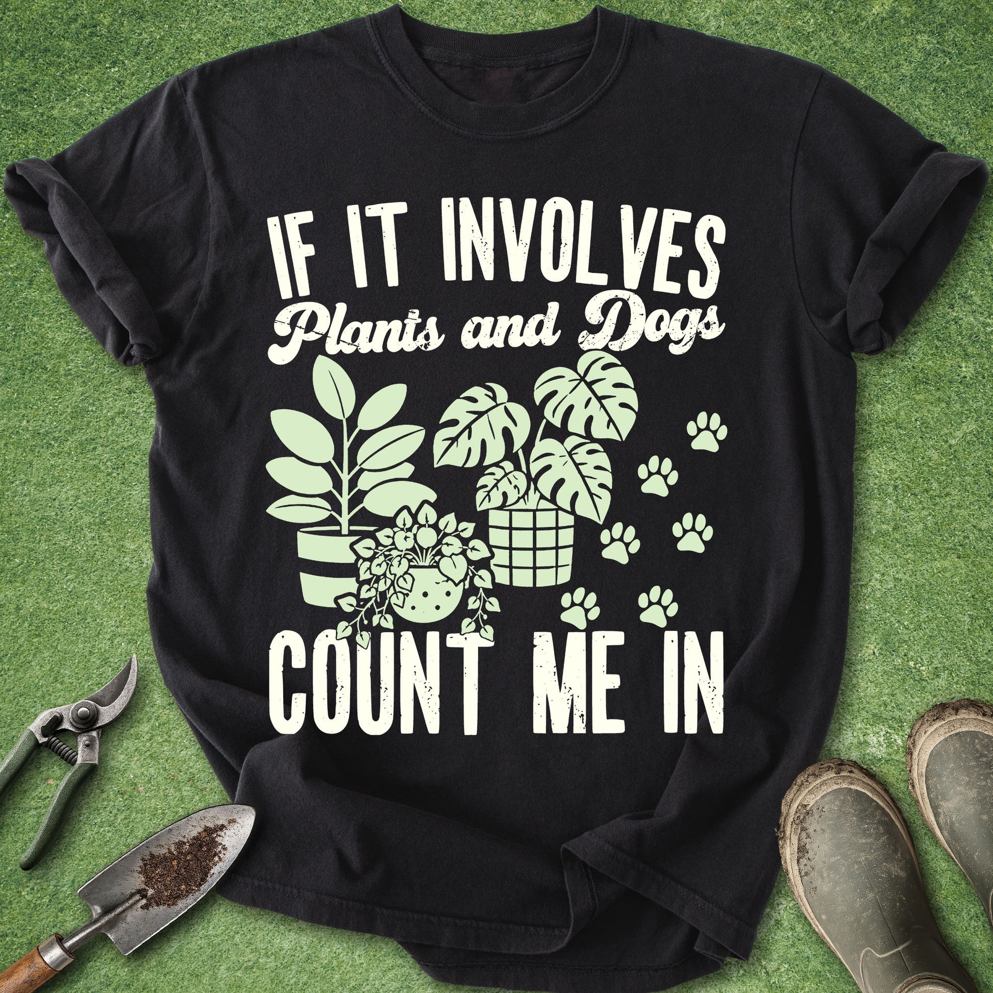 A black short-sleeve graphic T-shirt laid flat on green grass-like ground, with gardening tools and boots around it. The shirt has large white text reading 'IF IT INVOLVES Plants and Dogs COUNT ME IN' and a light green illustration of potted plants, leaves, and paw prints.