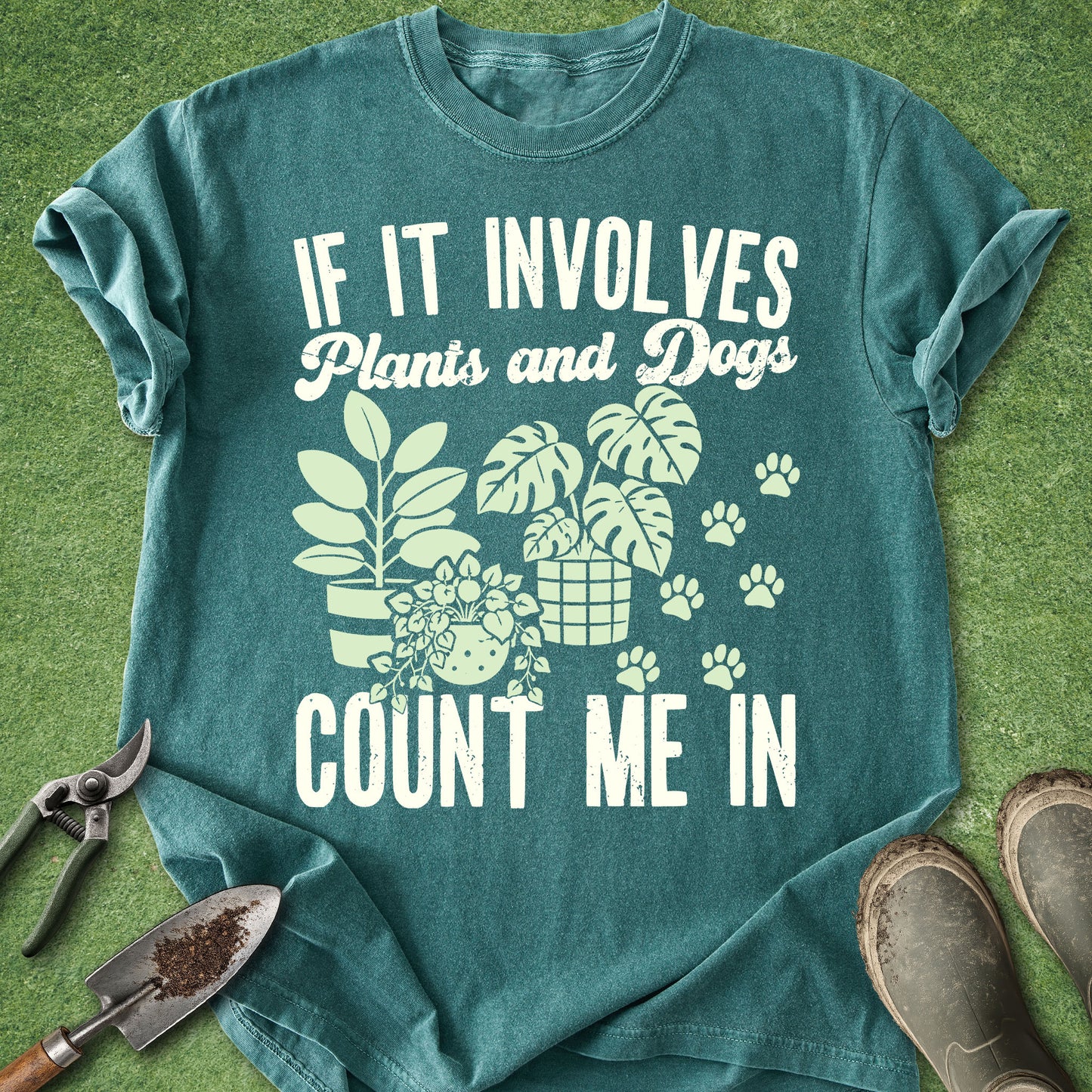 A teal graphic t-shirt laid flat on green grass, featuring the white printed text 'IF IT INVOLVES Plants and Dogs COUNT ME IN' along with illustrations of potted plants, a dog, and paw prints. Gardening tools and boots are placed around the shirt.