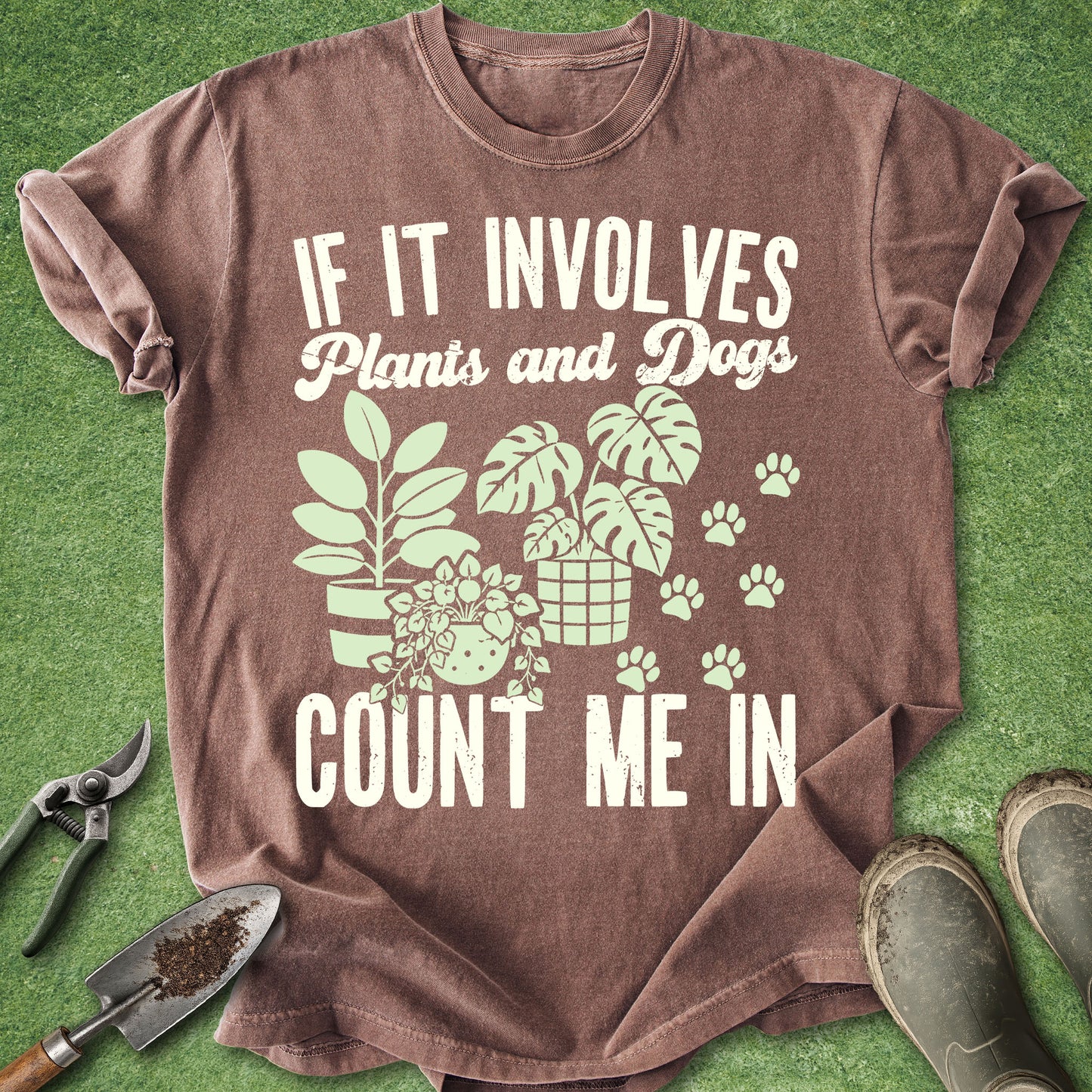 A brown short-sleeve graphic T-shirt laid flat on green artificial grass, with white text reading 'IF IT INVOLVES Plants and Dogs COUNT ME IN' and a plant-and-dog illustration. Gardening tools and boots are placed around the shirt as styling props.