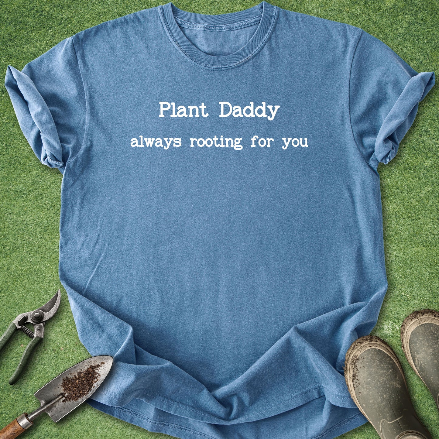 A blue crewneck t-shirt with the white printed text 'Plant Daddy always rooting for you' laid flat on green grass-like artificial turf, styled with gardening tools (trowel, pruning shears) and gardening boots.
