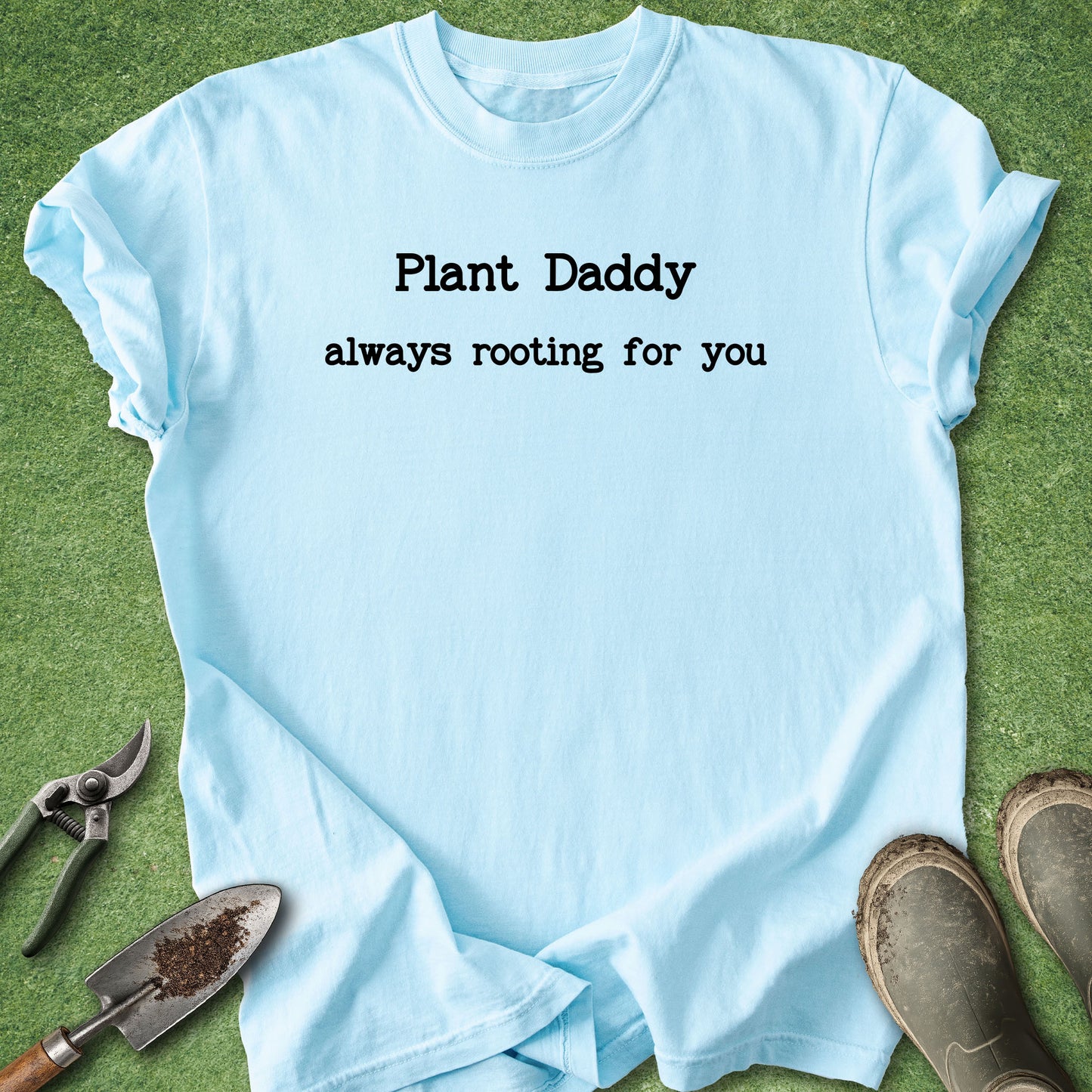 A light blue graphic t-shirt laid flat on green grass with gardening tools and boots around it. The t-shirt features the black printed text 'Plant Daddy always rooting for you'.