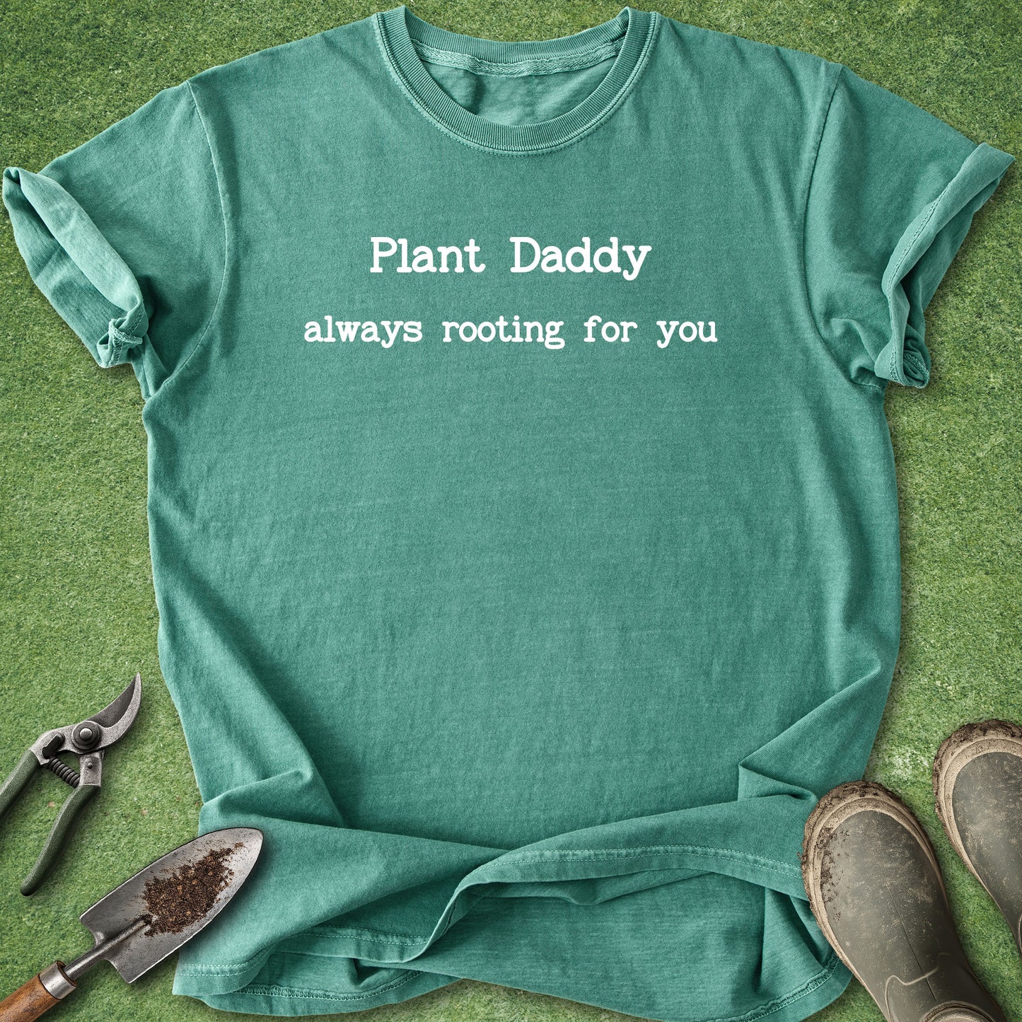 A teal green graphic t-shirt laid flat on green grass, featuring the white printed text 'Plant Daddy always rooting for you'. Gardening props, including a trowel, pruning shears, and boots, are arranged around the shirt.