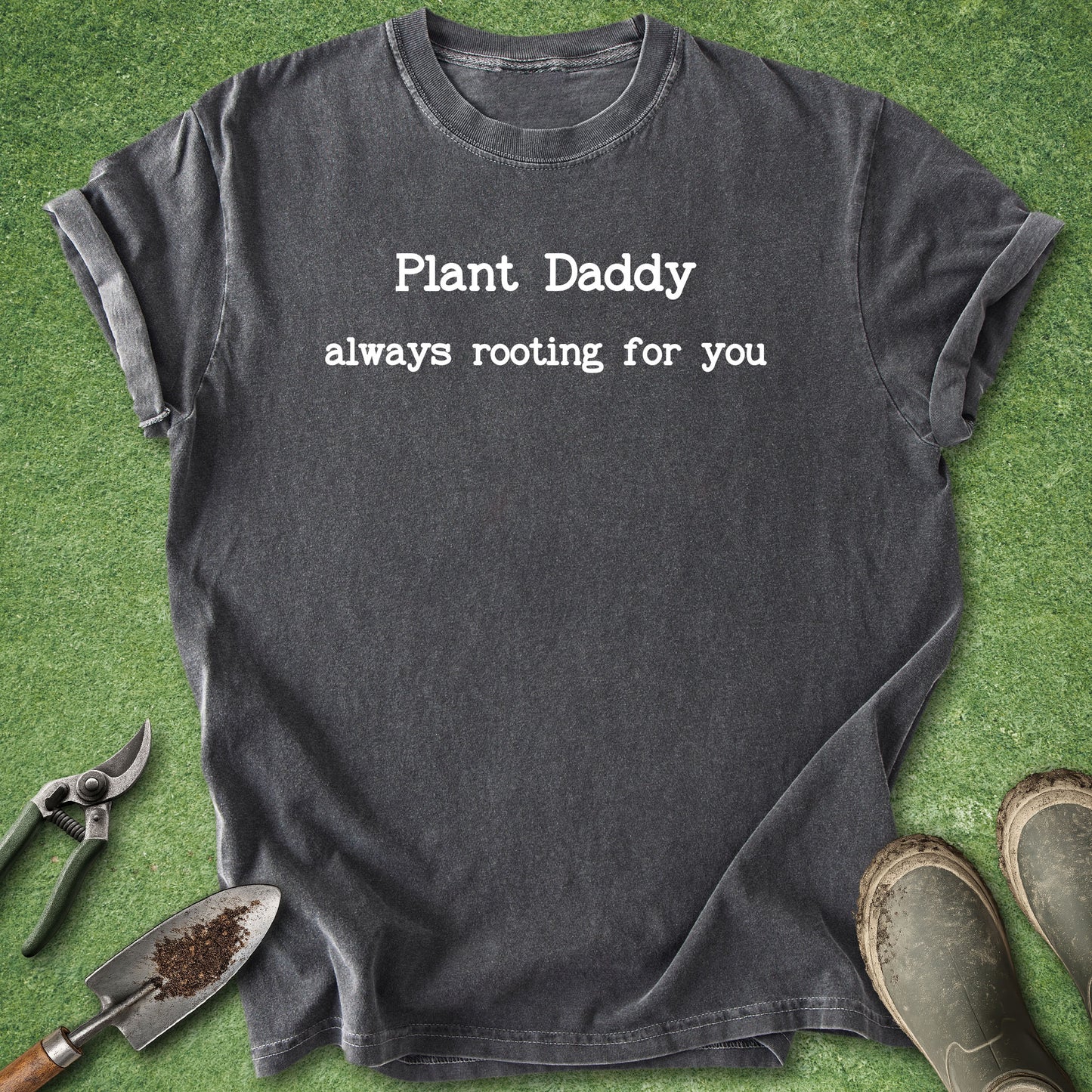 A dark charcoal gray graphic t-shirt with the text 'Plant Daddy always rooting for you' printed in white. The t-shirt is laid flat on green grass with gardening props, including a trowel, pruning shears, and boots.