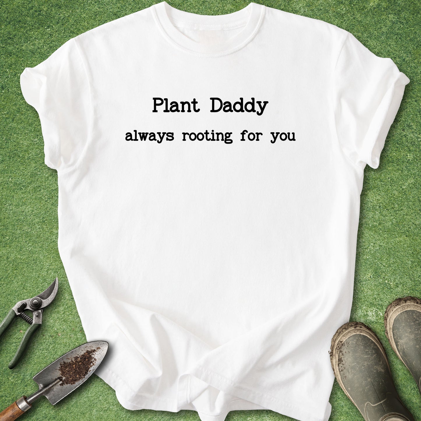 A white short-sleeve T-shirt laid flat on green grass-like artificial turf, with black text reading 'Plant Daddy always rooting for you.' Gardening props including pruning shears, a small trowel with soil, and muddy boots are placed around it.
