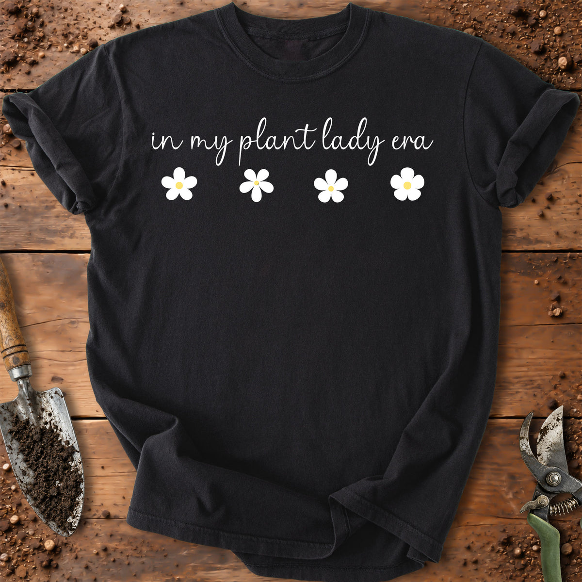 A black short-sleeve t-shirt with the text 'in my plant lady era' and four white daisy graphics, laid flat on a rustic wooden surface with gardening tools nearby.