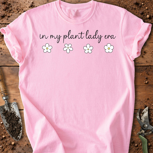 A light pink short-sleeve t-shirt laid flat on a rustic wooden surface with gardening tools. The shirt features the text 'in my plant lady era' in black script, followed by four small white daisy flowers with yellow centers.