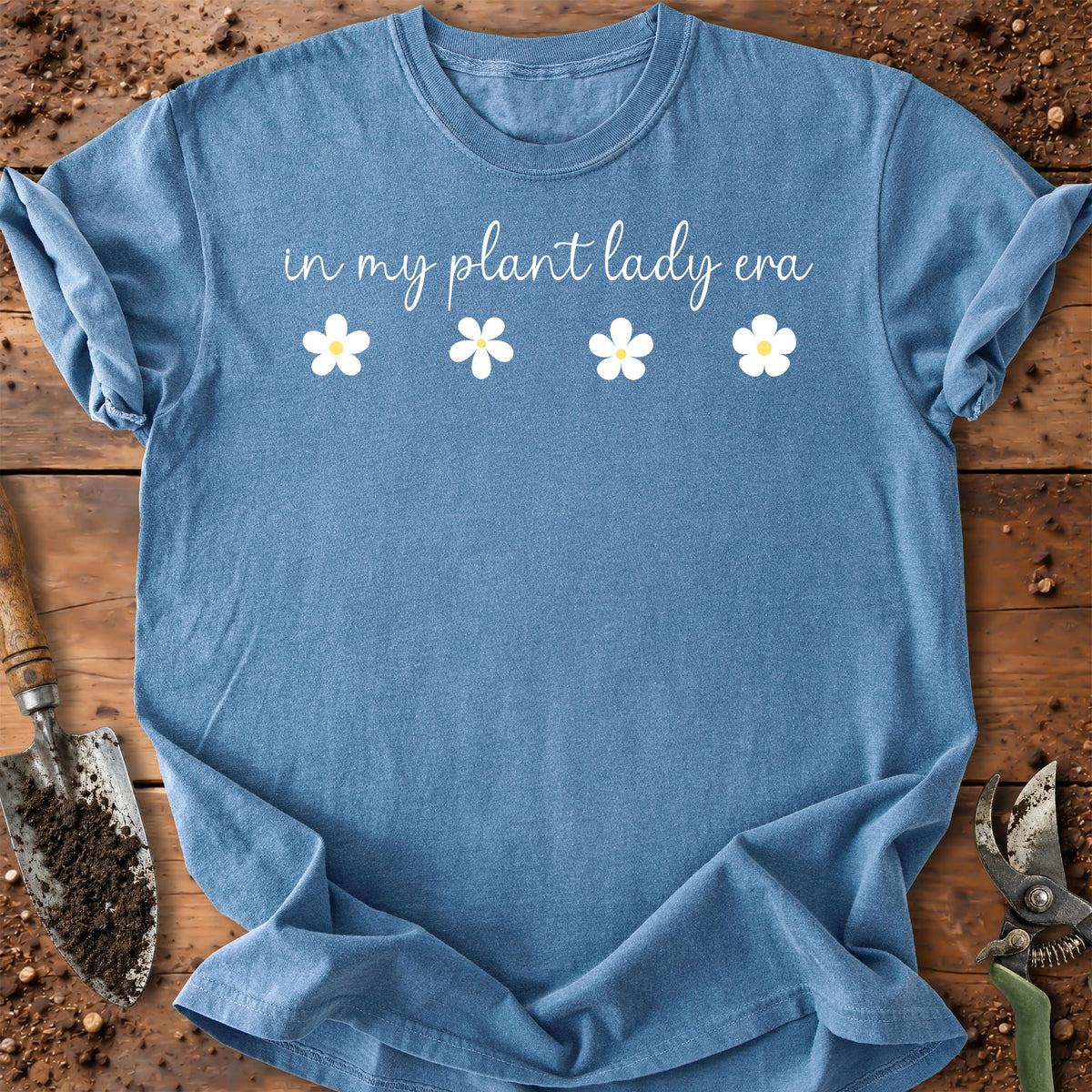 A blue short-sleeve t-shirt laid flat on a rustic wooden surface with gardening props like a trowel and pruning shears. The shirt features the white text 'in my plant lady era' above four small white daisy flowers.
