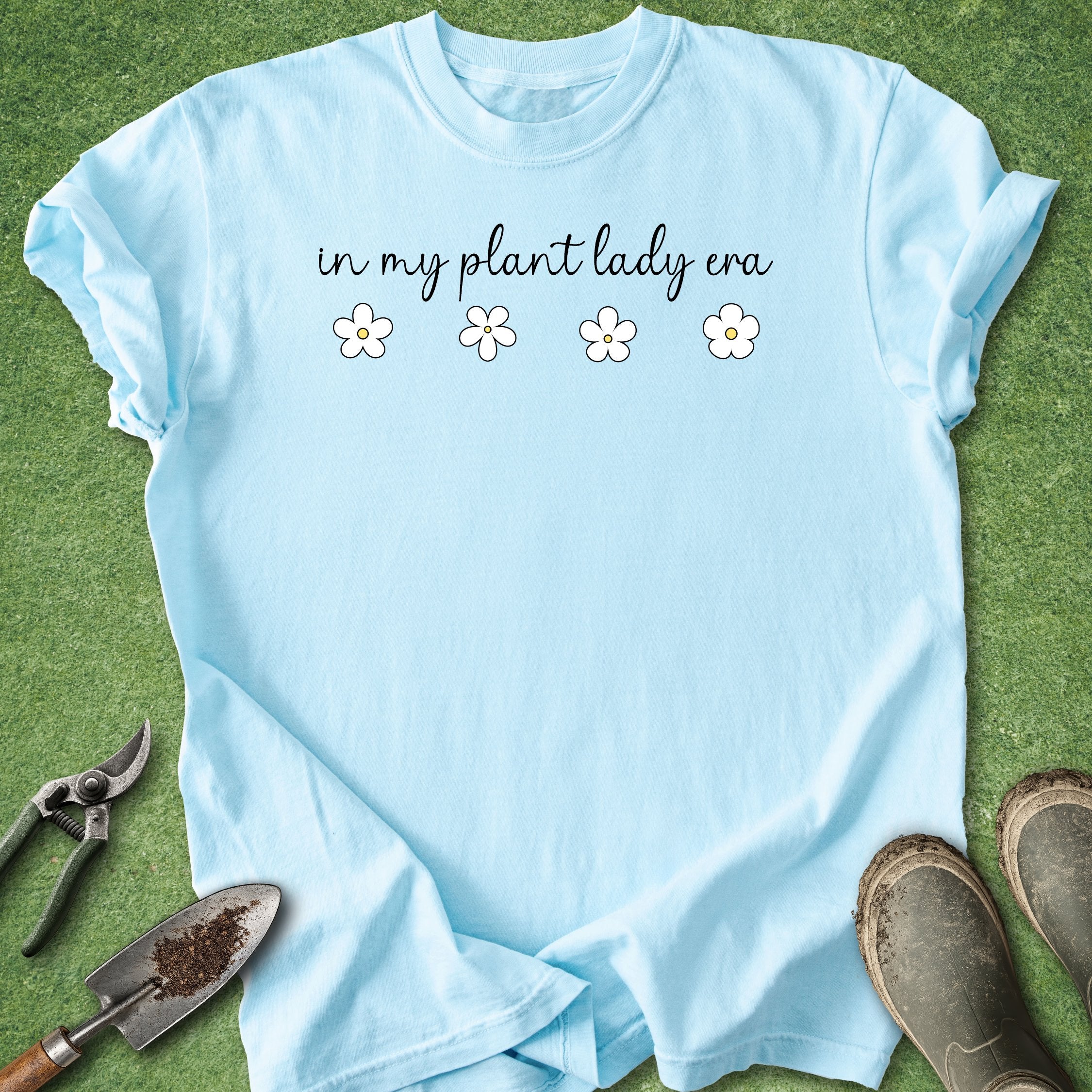 Light blue t-shirt with 'in my plant lady era' text and flowers on a grass background with gardening tools.