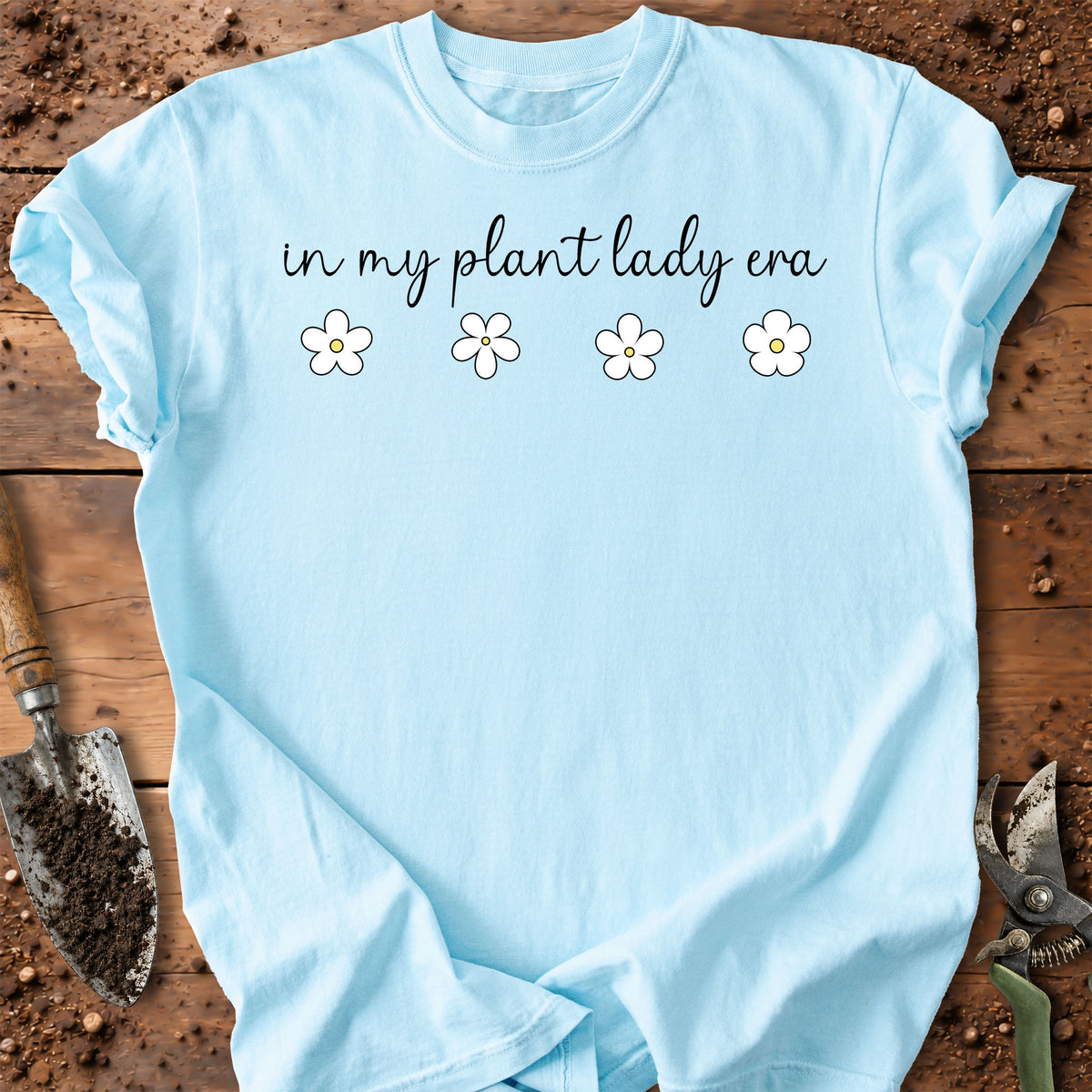 A light blue crewneck t-shirt with the text 'in my plant lady era' and four small white flowers printed on the chest, styled in a flat-lay on a rustic wooden surface with gardening tools.
