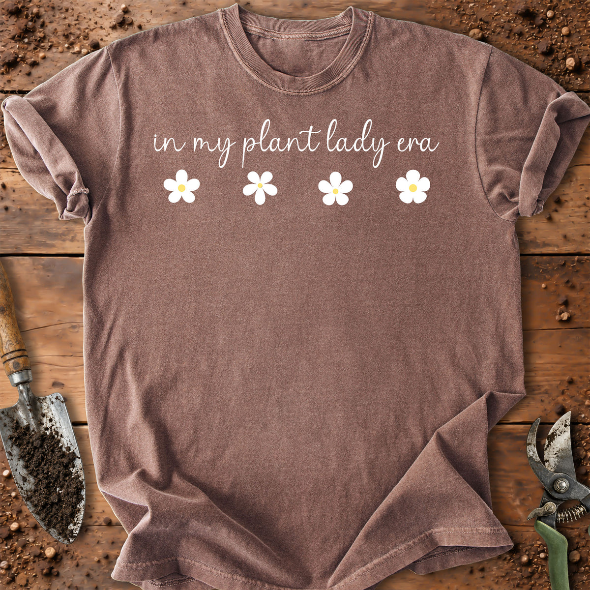 A brown short-sleeve t-shirt with the text 'in my plant lady era' and four white flowers printed on the chest, laid flat on a rustic wooden surface with gardening tools.