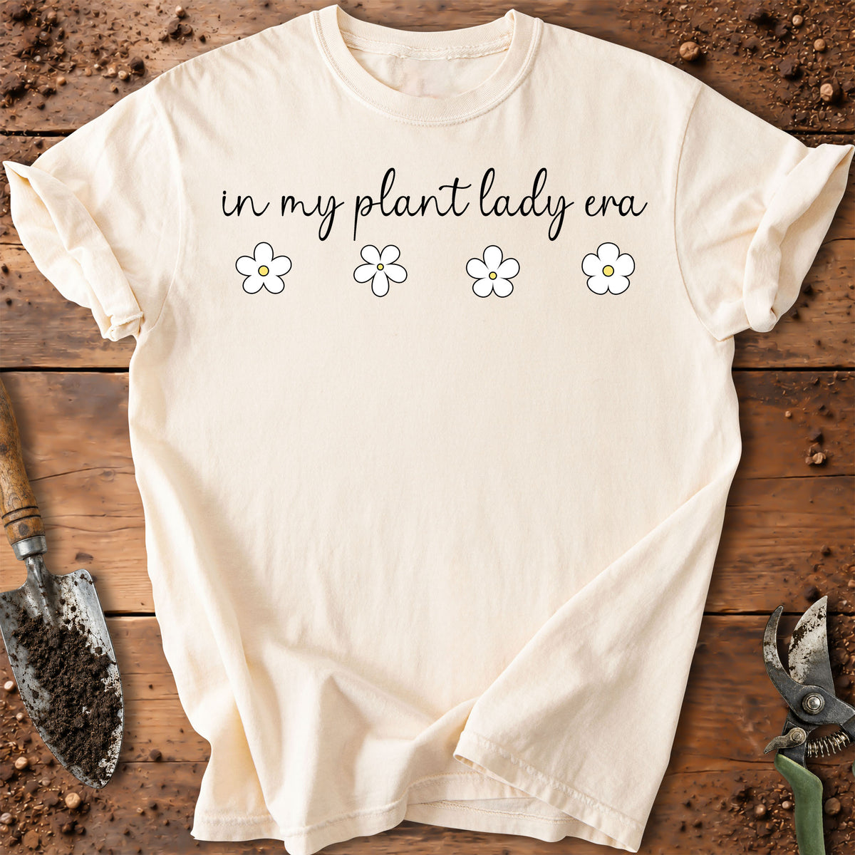 An off-white t-shirt with a black graphic reading 'dirt nerd' above simple line drawings of a shovel, rake, and pruning shears, laid flat on a rustic wooden surface with gardening tools around it.