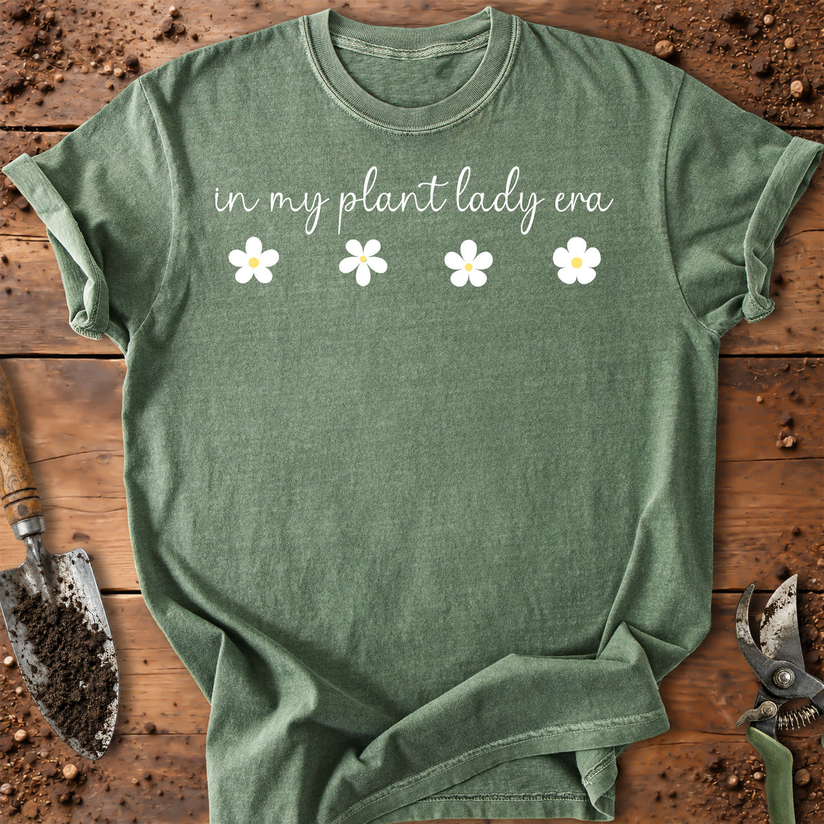 An olive green short-sleeve t-shirt with the text 'in my plant lady era' and four white daisy graphics, laid flat on a rustic wooden surface with gardening tools.