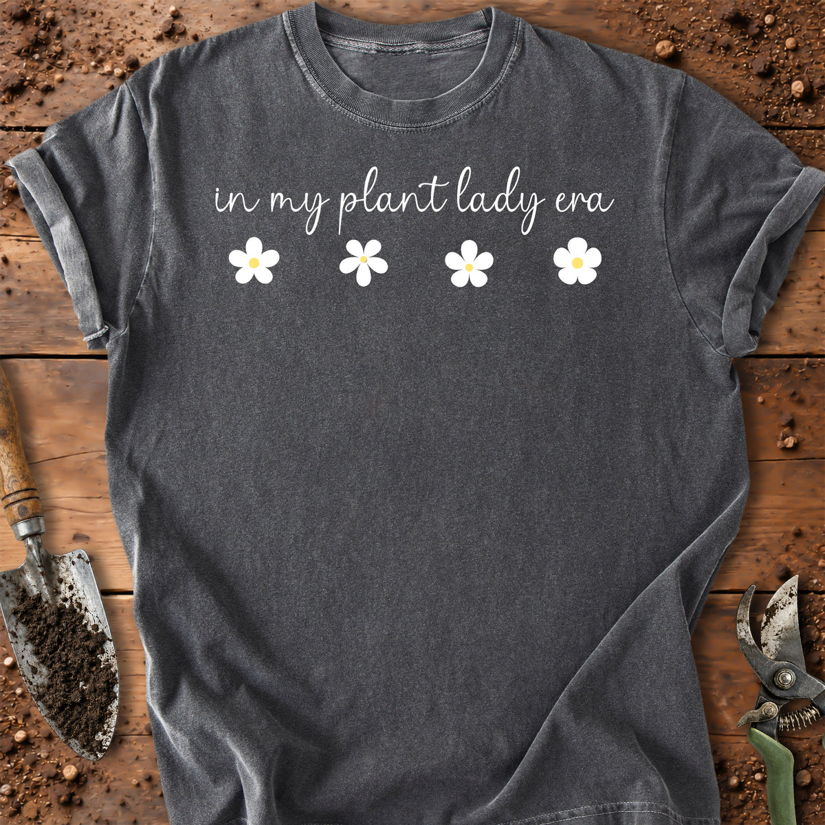 A dark gray t-shirt with the text 'in my plant lady era' and four white daisy graphics, laid flat on a rustic wooden surface with gardening tools.