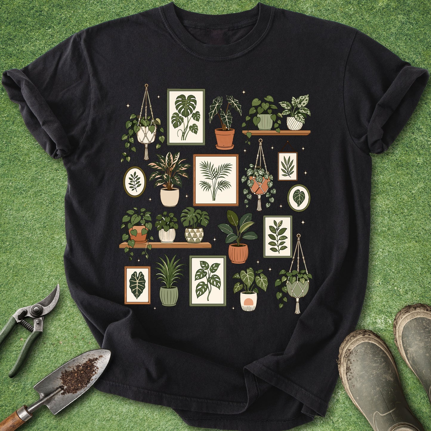 A black short-sleeve t-shirt laid flat on green grass-like artificial turf, featuring a large central graphic print of various illustrated potted houseplants in frames and hanging macrame holders. Gardening props including pruning shears, a small hand trowel with soil, and work boots are placed around the shirt.