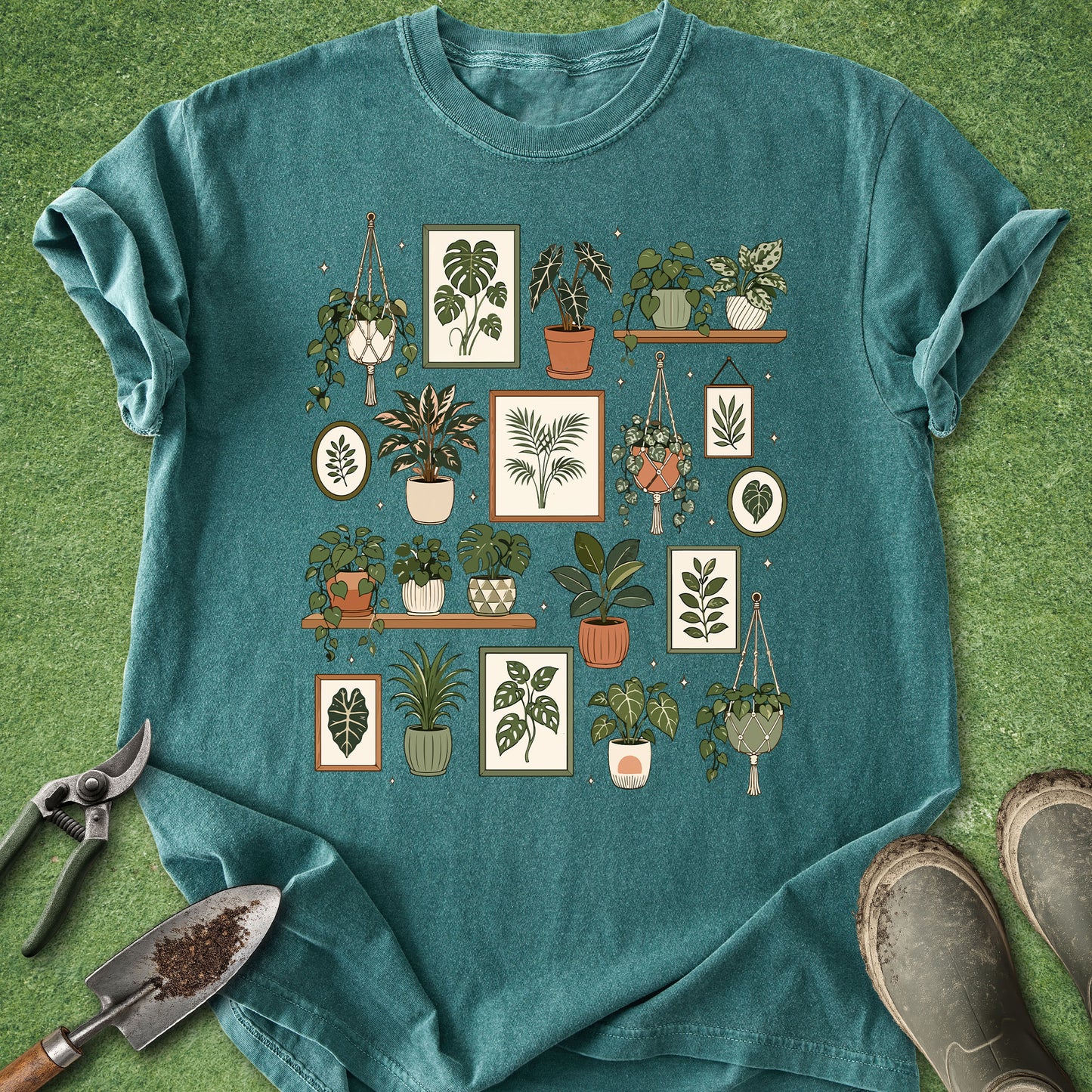 A teal short-sleeve t-shirt with a graphic print of various illustrated houseplants, shown laid flat on green grass with gardening tools and boots nearby.