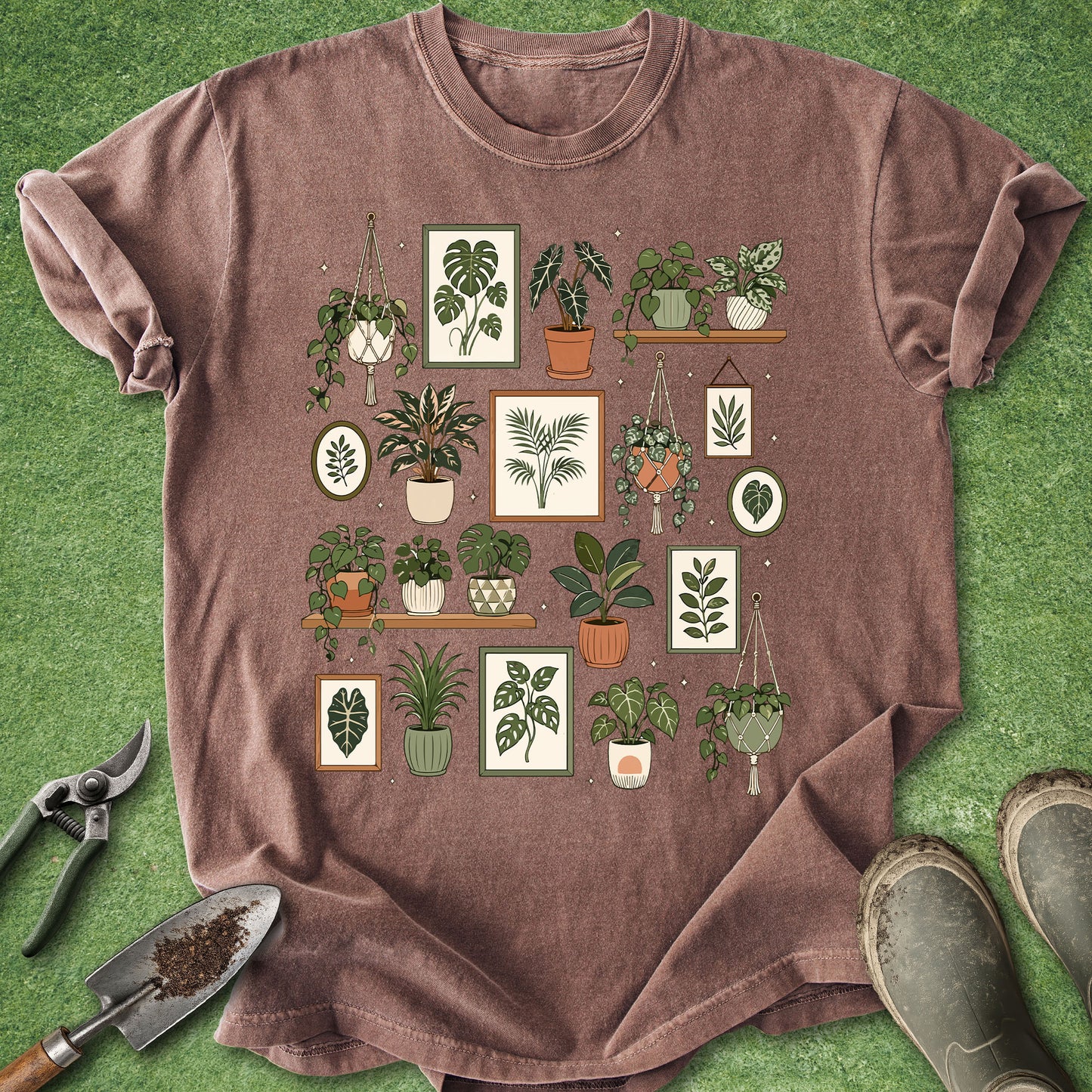 A heathered brown short-sleeve t-shirt with a graphic print of various potted houseplants, styled on a green grass-like surface with gardening tools and boots.