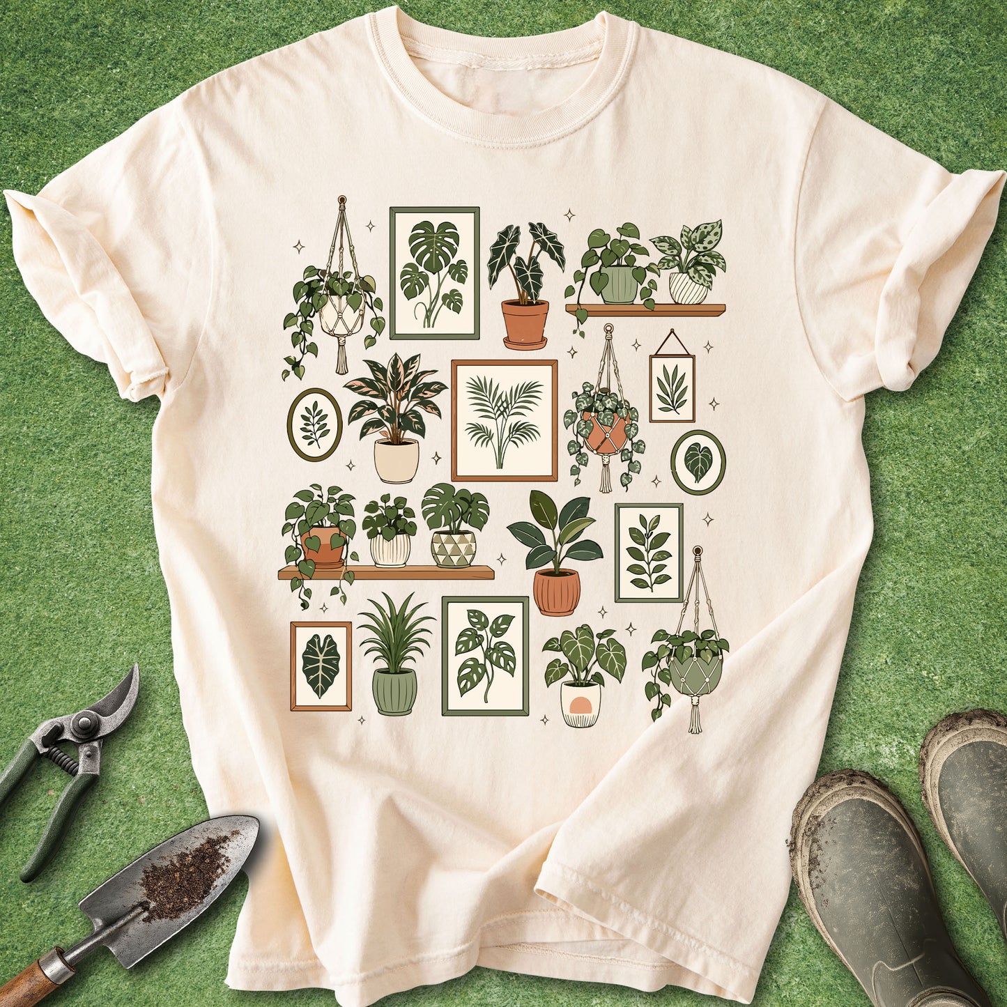A cream-colored short-sleeve t-shirt with a graphic print of various potted houseplants in frames, laid flat on a green grass-like surface with gardening tools and boots nearby.