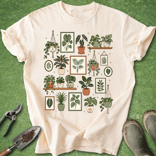 A cream-colored short-sleeve t-shirt with a graphic print of various potted houseplants in frames, laid flat on a green grass-like surface with gardening tools and boots nearby.
