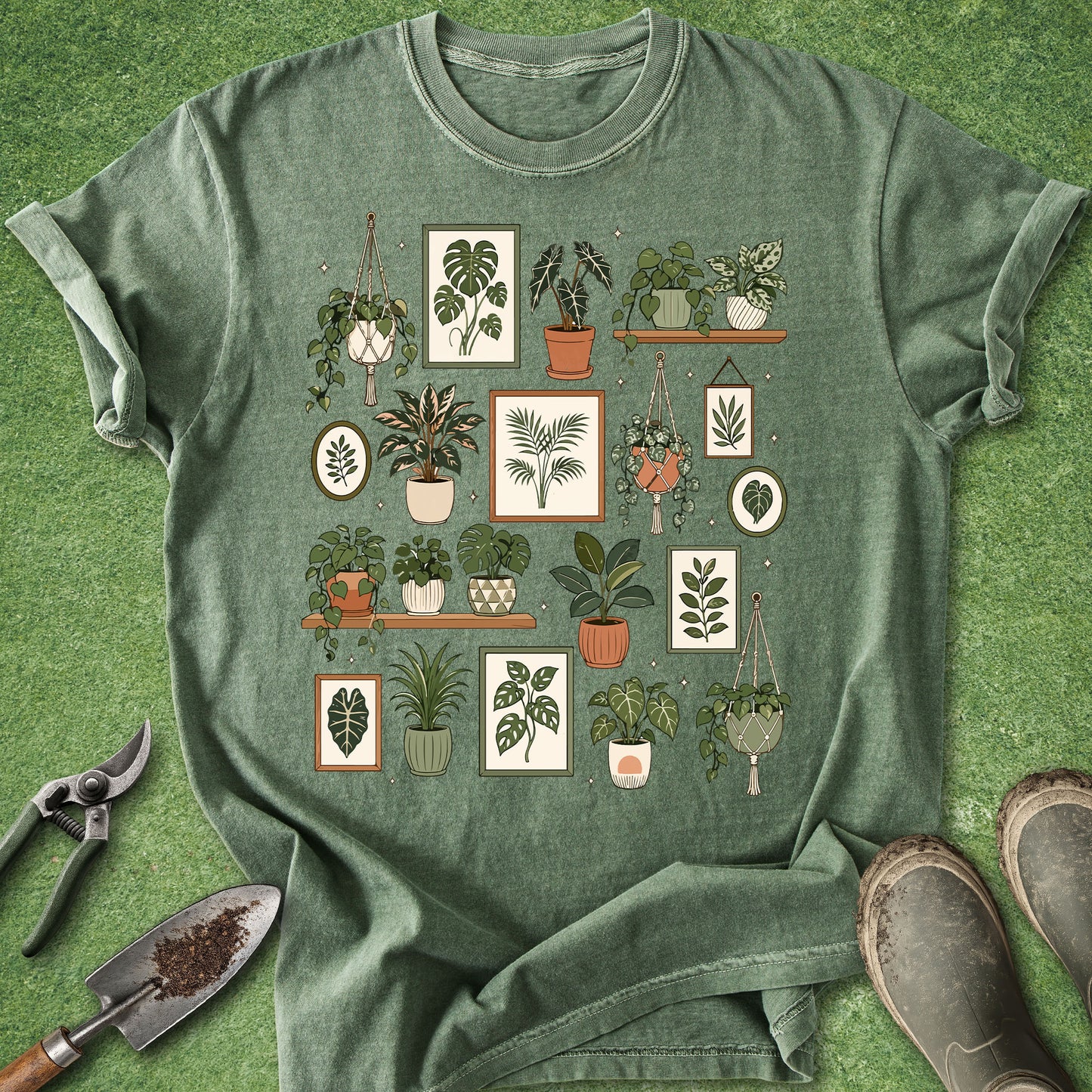 A sage green short-sleeve t-shirt with a graphic print of various potted houseplants, styled in a flat lay on artificial green grass with gardening tools (trowel, pruning shears) and muddy boots.