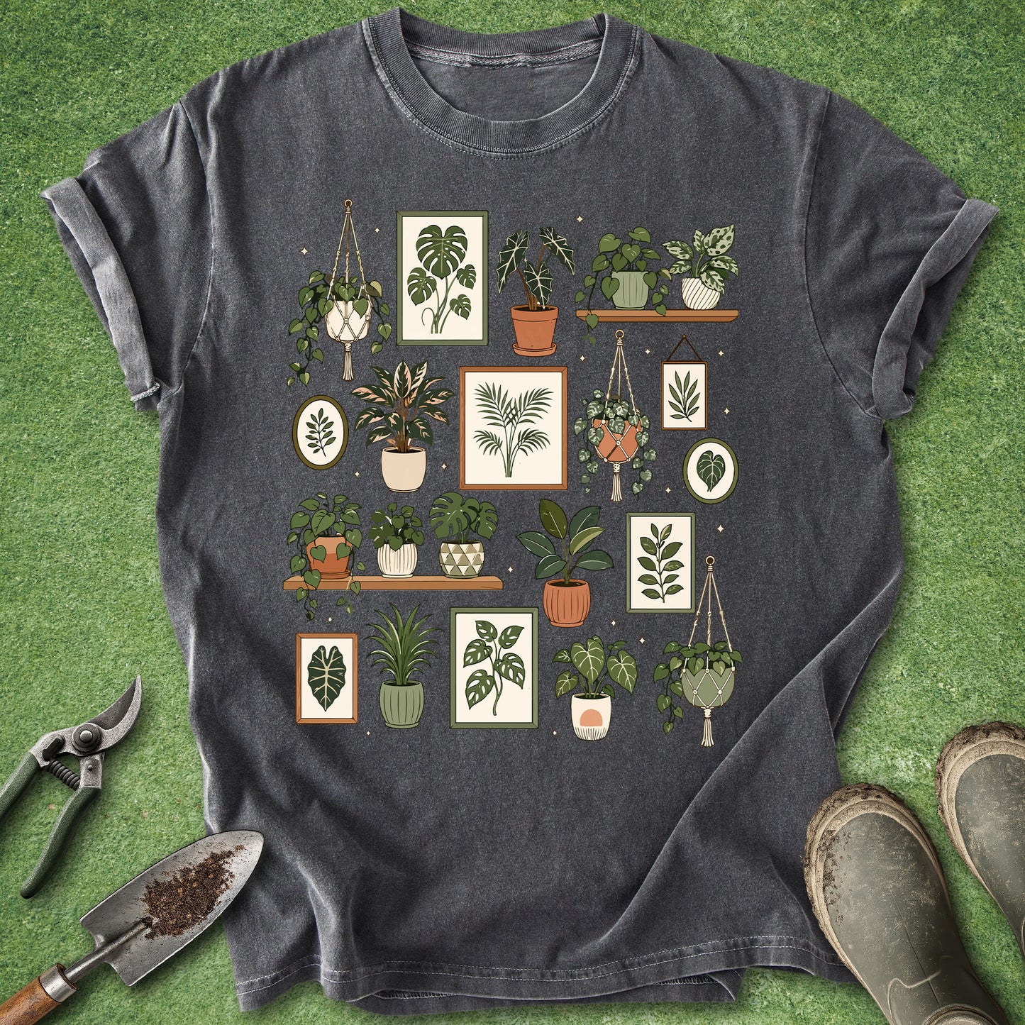 A dark gray graphic t-shirt featuring a grid of various potted houseplants and botanical prints, styled in a flat lay on artificial green grass with gardening tools (trowel, shears) and boots.