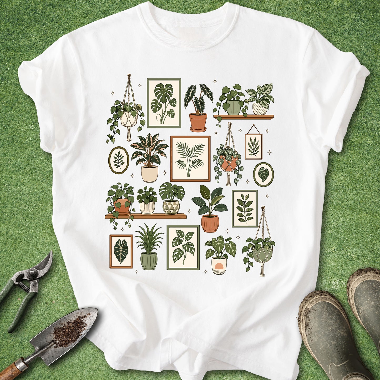 A white short-sleeve T-shirt laid flat on green grass, featuring a central graphic print of various illustrated potted houseplants in muted green, beige, and terracotta tones. Gardening tools and muddy boots are placed around the shirt as styling props.