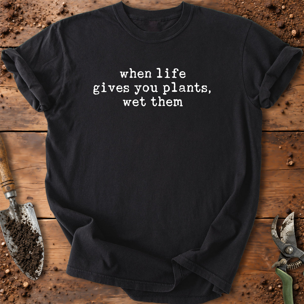 A black short-sleeve crewneck t-shirt with the white printed text 'when life gives you plants, wet them' laid flat on a rustic wooden surface with gardening props like a trowel and pruning shears.