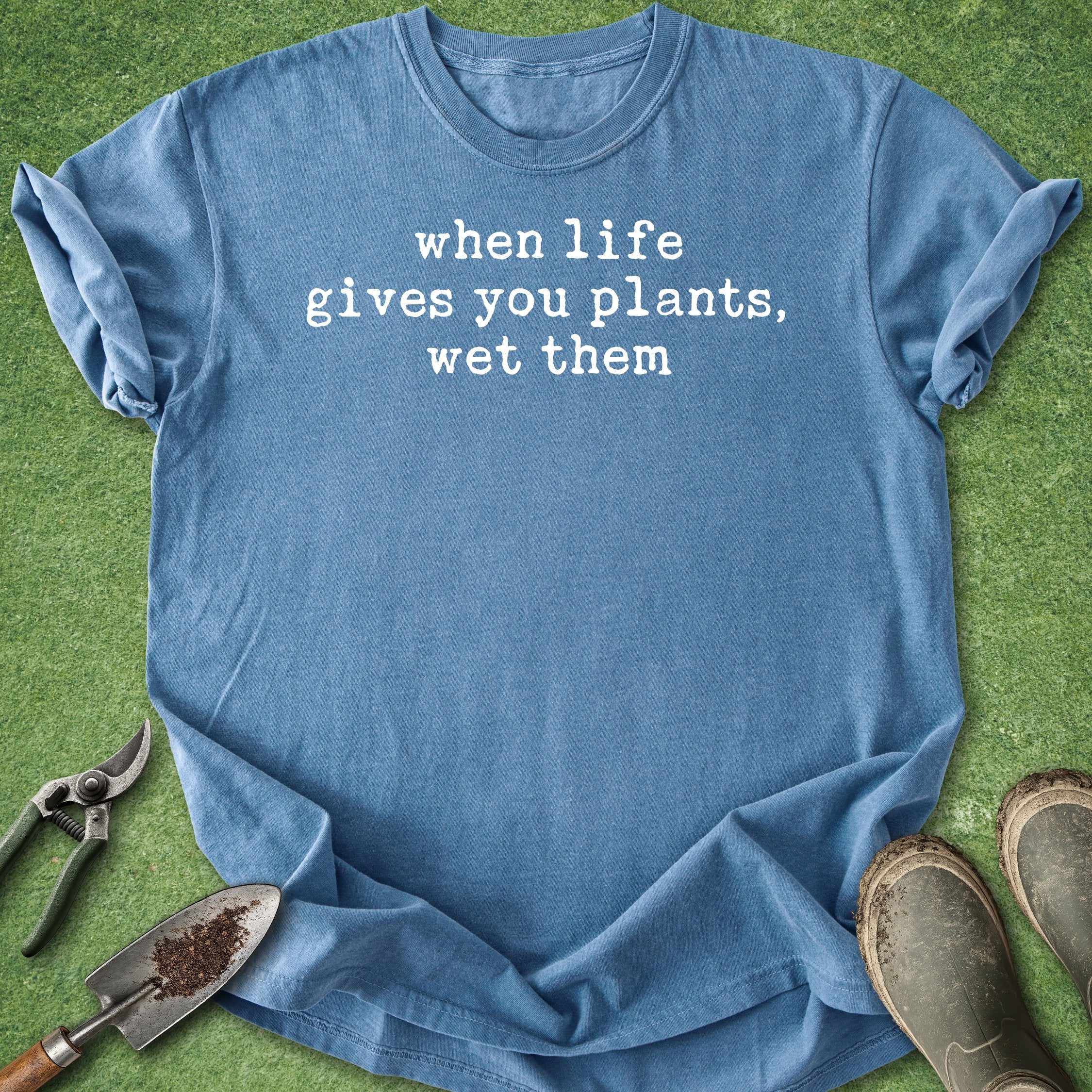 Blue t-shirt with 'when life gives you plants, wet them' text on a grass background with gardening tools.