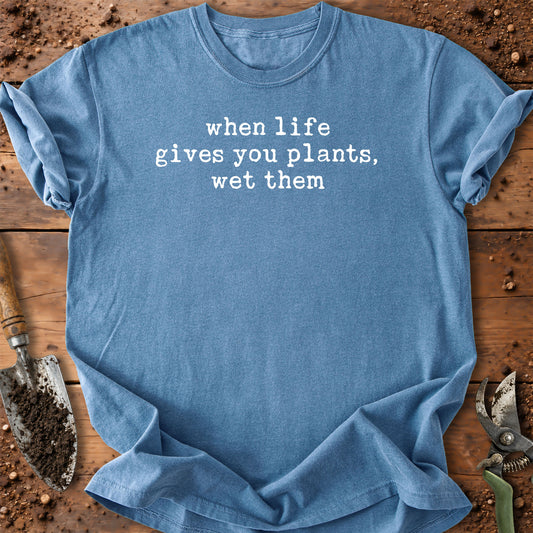 A blue graphic t-shirt with the text 'when life gives you plants, wet them' in white typewriter font, laid flat on a rustic wooden surface with gardening tools like a trowel and pruning shears.