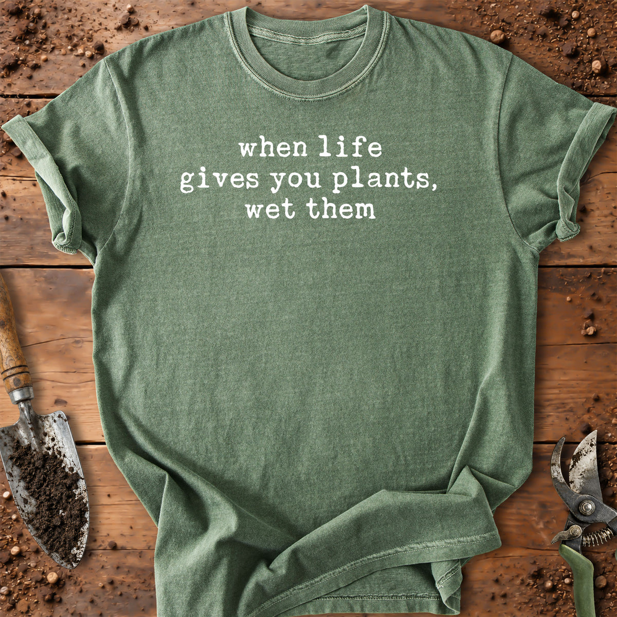 An olive green crewneck t-shirt with the white printed text 'when life gives you plants, wet them' laid flat on a rustic wooden surface with gardening tools like a trowel and pruning shears.