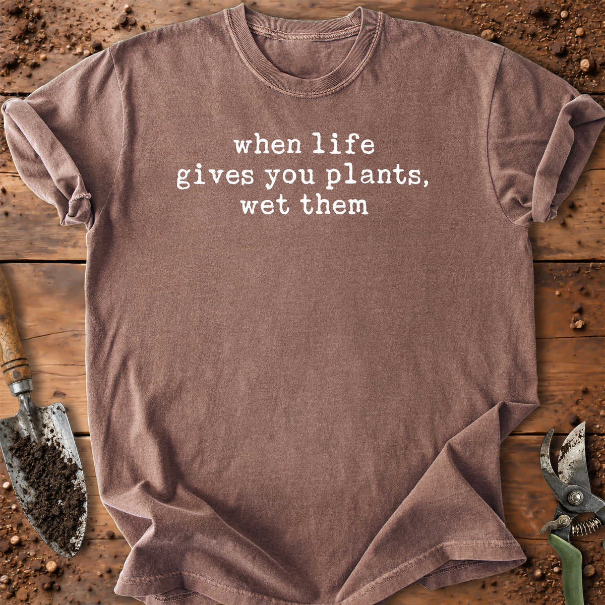 A heathered taupe-brown short-sleeve t-shirt with the phrase 'when life gives you plants, wet them' printed in white lowercase serif text across the chest. The shirt is laid flat on a rustic wooden surface surrounded by gardening props, including a trowel and pruning shears.