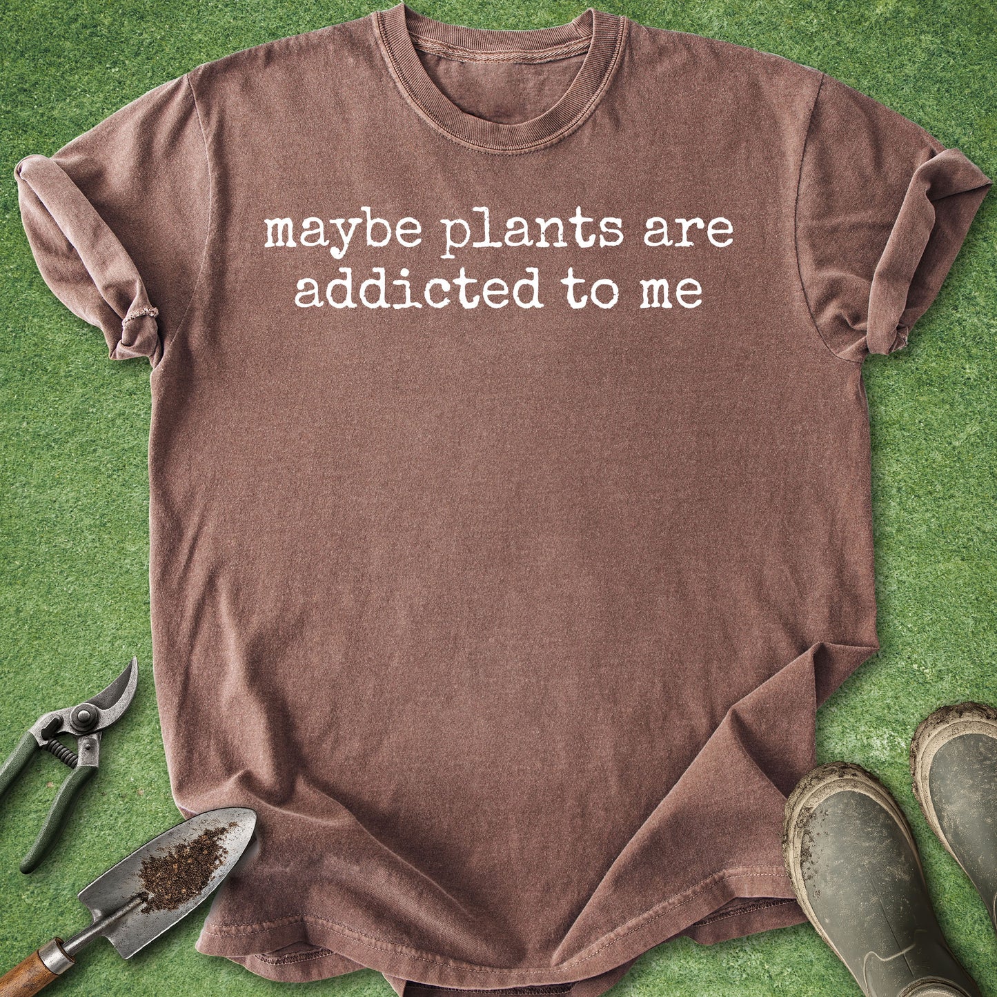 A brown short-sleeve t-shirt with the white text 'maybe plants are addicted to me' printed on the chest, laid flat on a green grass-like surface with gardening tools and boots nearby.