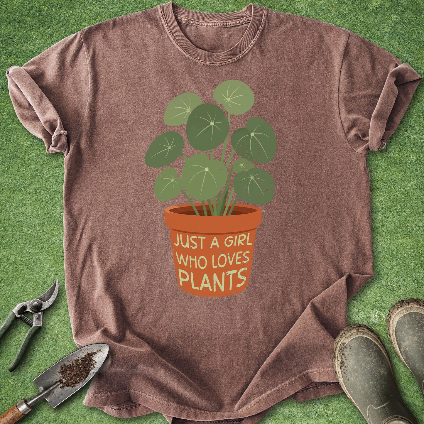 A heathered brown short-sleeve t-shirt laid flat on green grass, featuring a graphic of a potted plant with the text 'JUST A GIRL WHO LOVES PLANTS'. Gardening props including pruning shears, a small hand trowel with soil, and rubber boots are arranged around the shirt.