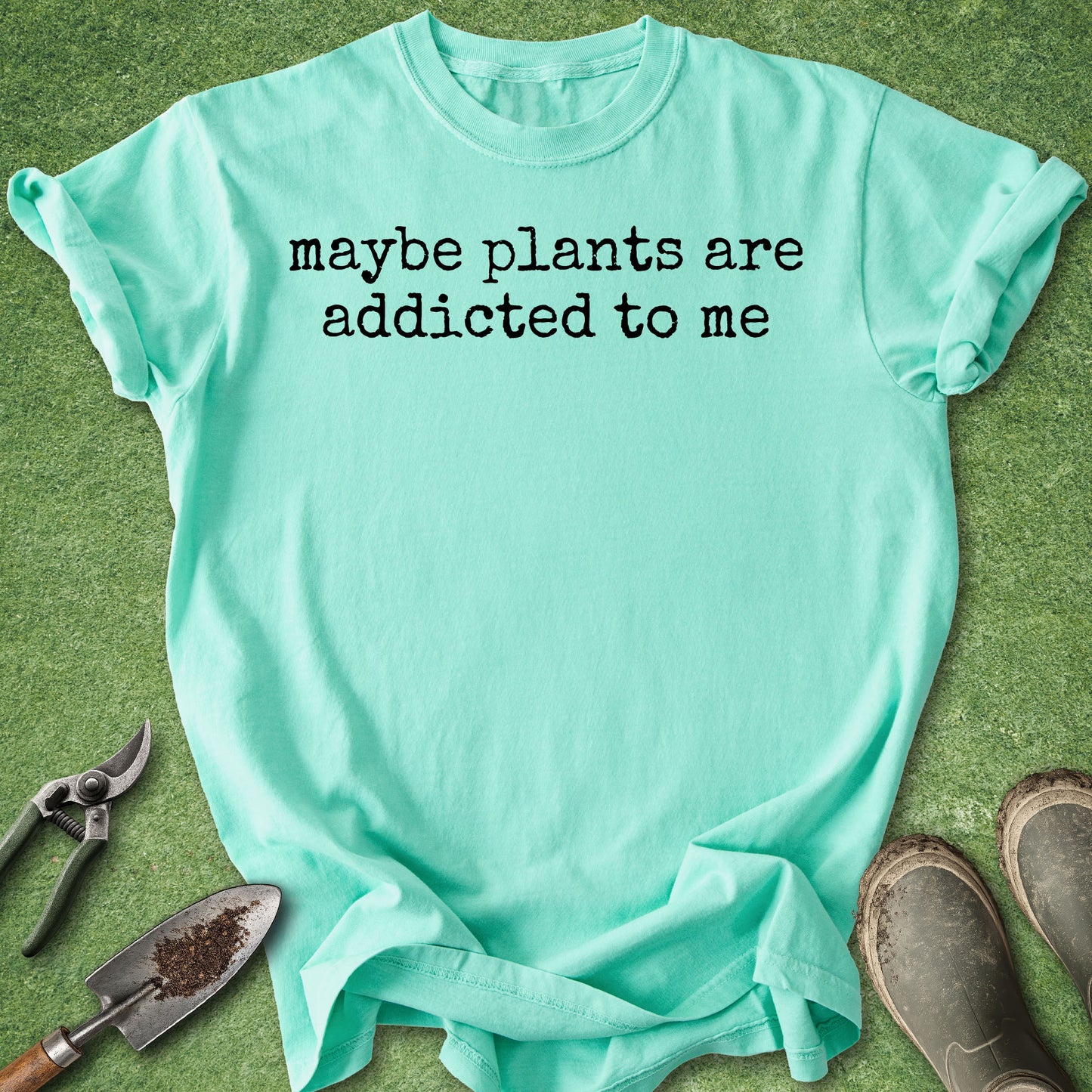 A mint green graphic t-shirt laid flat on green grass with gardening props, including pruning shears, a trowel, and work boots. The t-shirt features the black printed text 'maybe plants are addicted to me'.