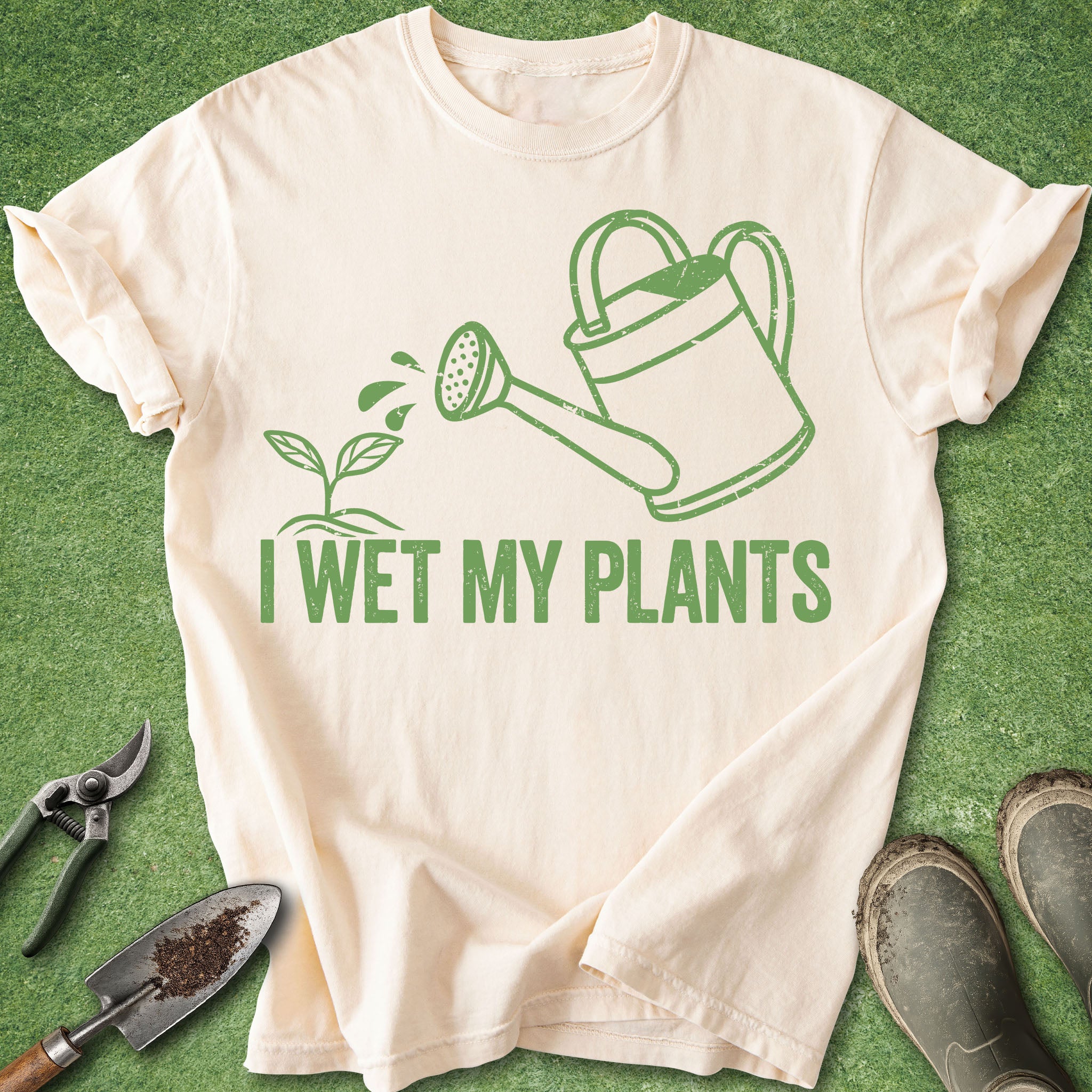 Beige t-shirt with green graphic and 'I Wet My Plants' text on a grass background