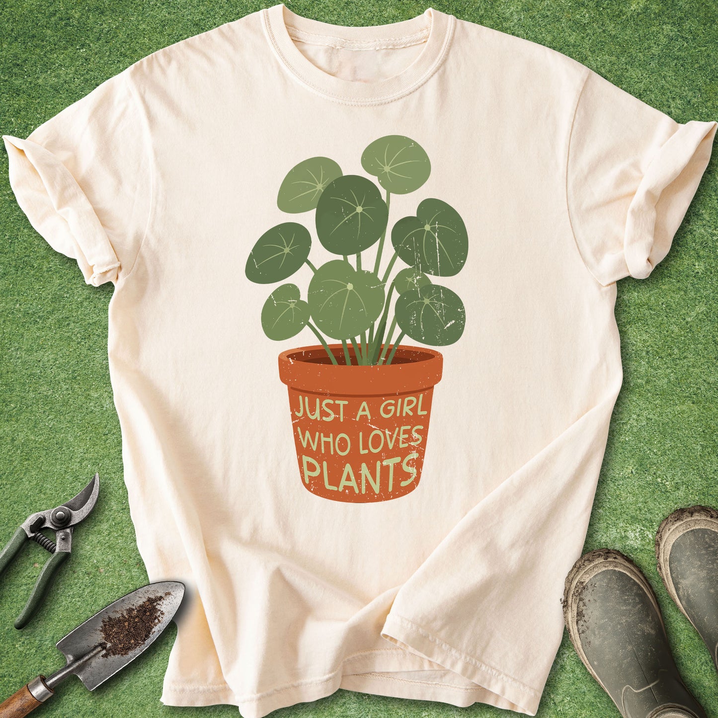 A cream-colored short-sleeve t-shirt with a graphic of a potted plant and the text 'JUST A GIRL WHO LOVES PLANTS', laid flat on a green grass-like background with gardening tools and shoes nearby.
