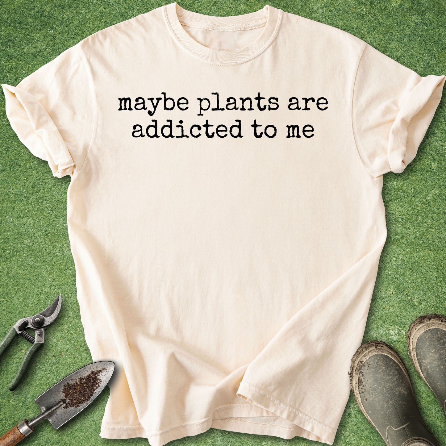 A cream-colored short-sleeve t-shirt with the black printed text 'maybe plants are addicted to me' laid flat on a green grass-like surface, styled with gardening tools and muddy boots.
