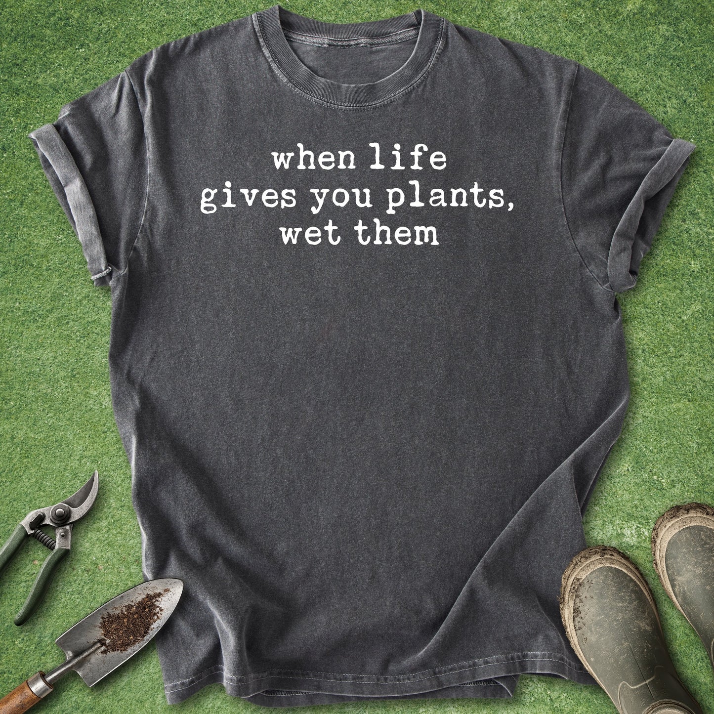 Black t-shirt with 'when life gives you plants, wet them' text on a grass background with gardening tools.