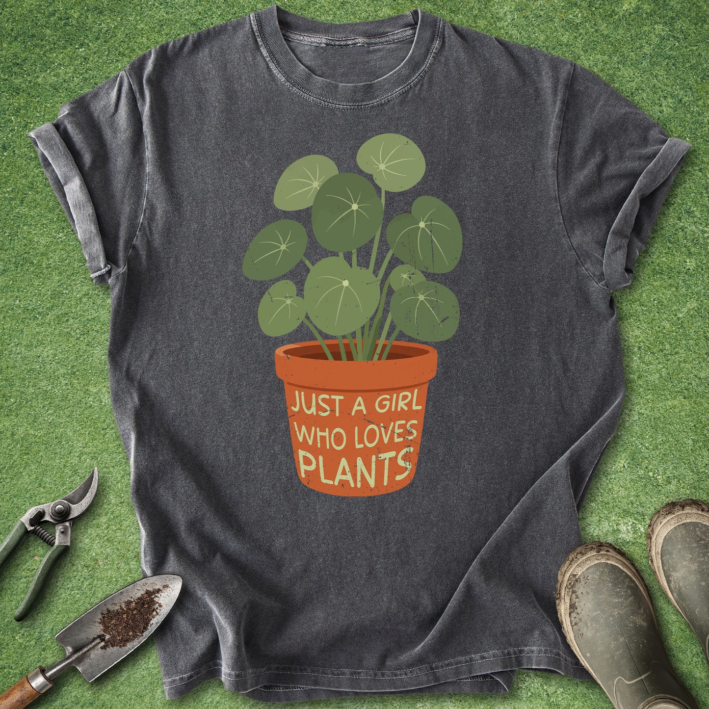 A dark gray graphic t-shirt laid flat on green grass, featuring a cartoon illustration of a potted plant with the text 'JUST A GIRL WHO LOVES PLANTS' on the pot. Gardening props, including pruning shears, a trowel, and boots, are arranged around the shirt.
