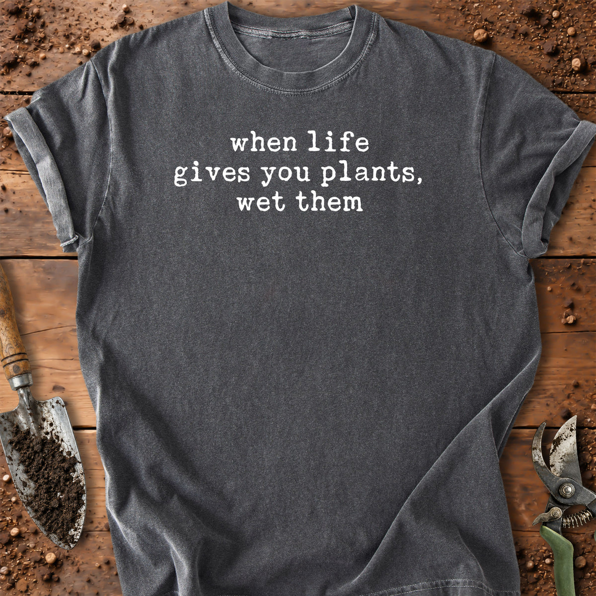 A dark gray t-shirt with the white printed text 'when life gives you plants, wet them' laid flat on a rustic wooden surface with gardening tools like a trowel and pruning shears.