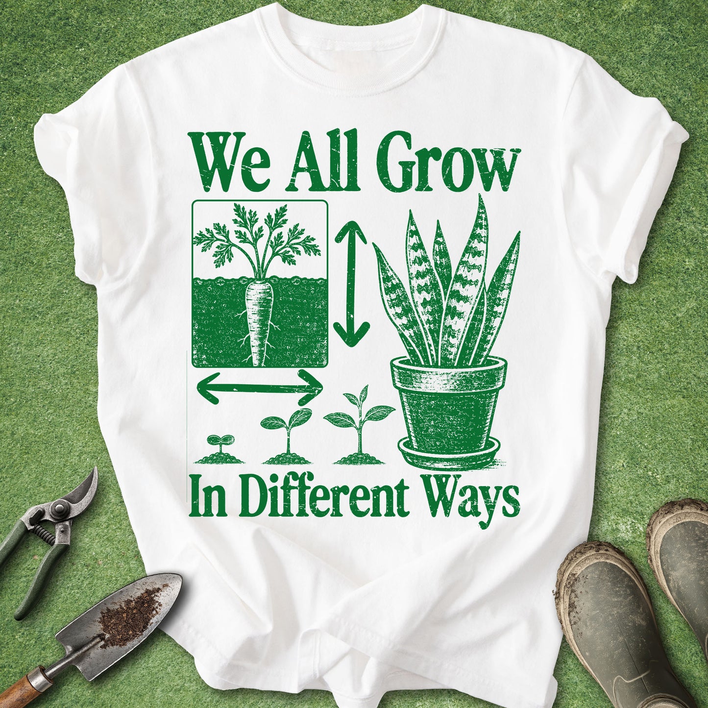 A white graphic T-shirt laid flat on green grass-like background, with gardening tools and boots around it. The shirt has a large green printed design reading 'We All Grow In Different Ways' with plant illustrations and growth arrows.
