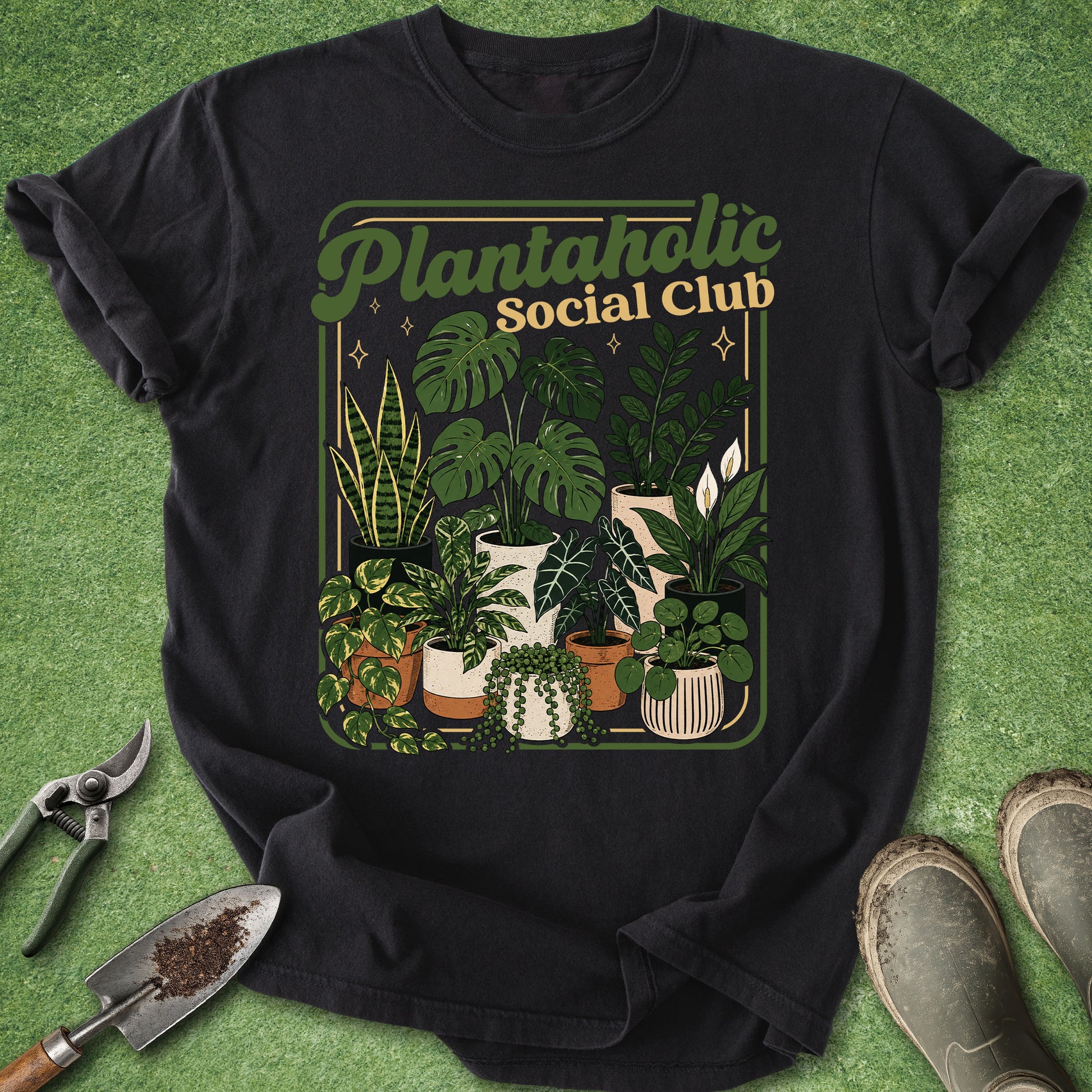 A black short-sleeve graphic T-shirt laid flat on a green textured surface, surrounded by gardening props including pruning shears, a trowel, and boots. The shirt features a large retro-style plant illustration with the text 'Plantaholic Social Club' across the chest.