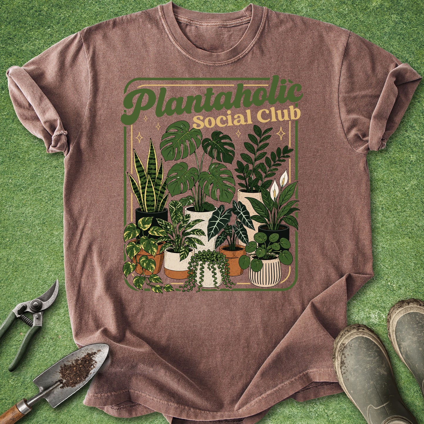 A brown short-sleeve graphic T-shirt laid flat on a green textured surface, surrounded by gardening props including pruning shears, a trowel, and boots. The shirt features a retro-style 'Plantaholic Social Club' print with illustrated potted plants.