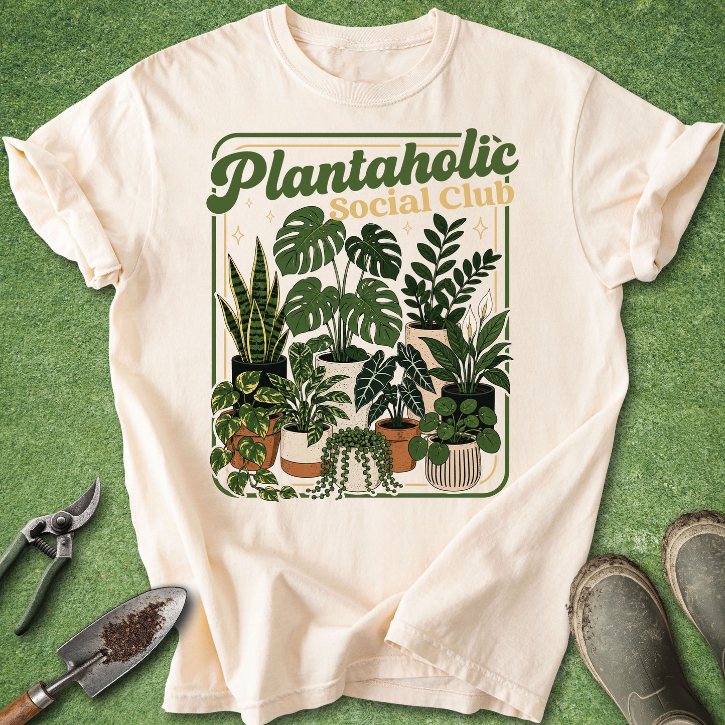 An off-white short-sleeve t-shirt with a retro-style 'Plantaholic Social Club' graphic featuring various green potted plants, laid flat on green artificial turf with gardening tools and boots nearby.