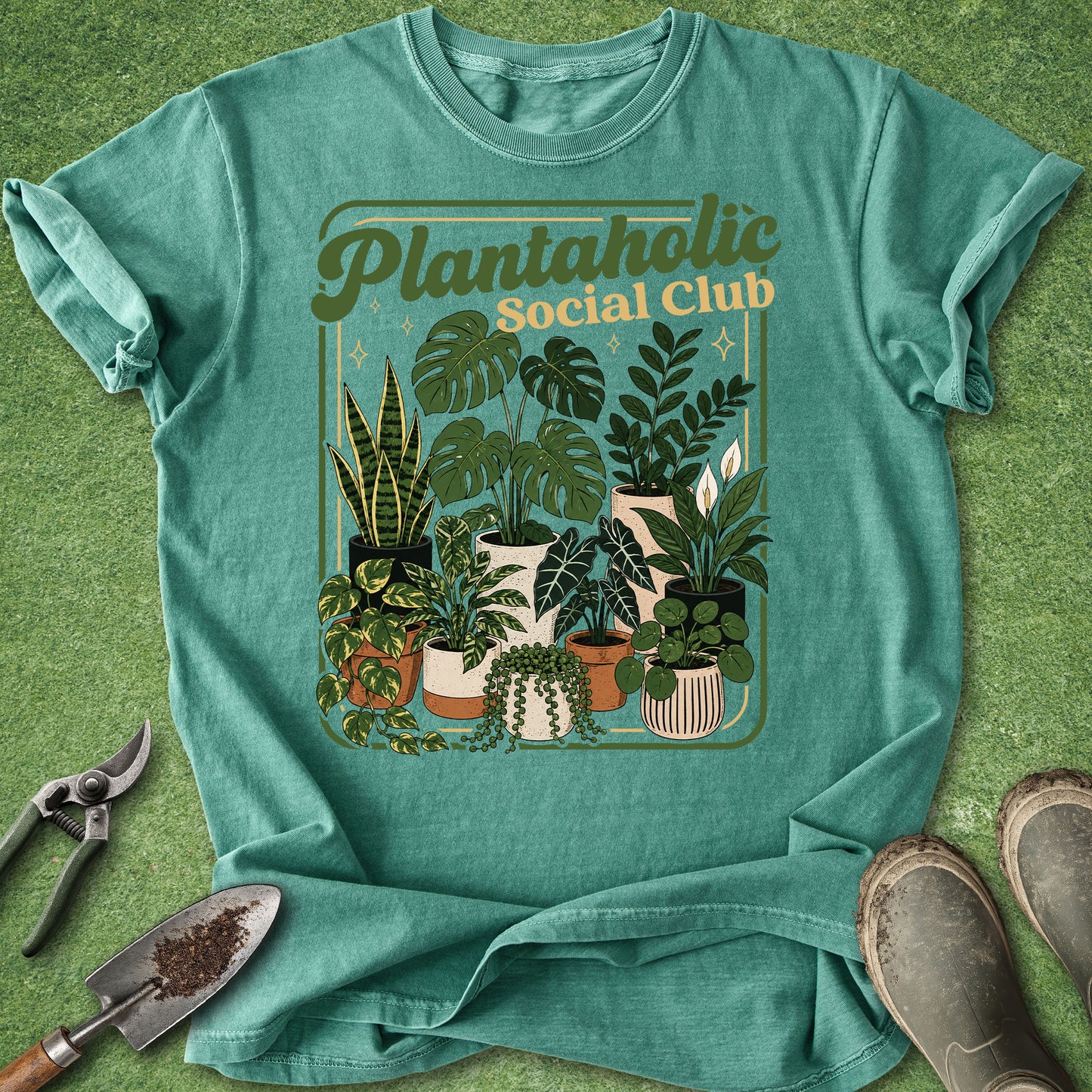 A teal short-sleeve graphic T-shirt laid flat on a green textured surface, featuring a retro-style illustration of potted plants and the words 'Plantaholic Social Club.' Gardening tools and boots are placed around the shirt as styling props.