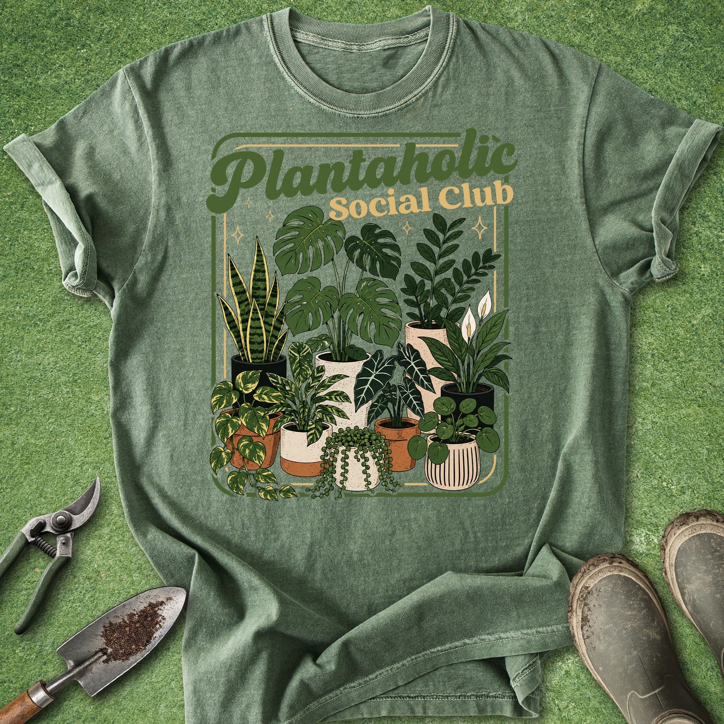 A sage green graphic t-shirt laid flat on a green grass-like surface, featuring a retro-style illustration of various potted plants with the text 'Plantaholic Social Club' in olive green and tan. Gardening props including pruning shears, a trowel, and boots are arranged around the shirt.