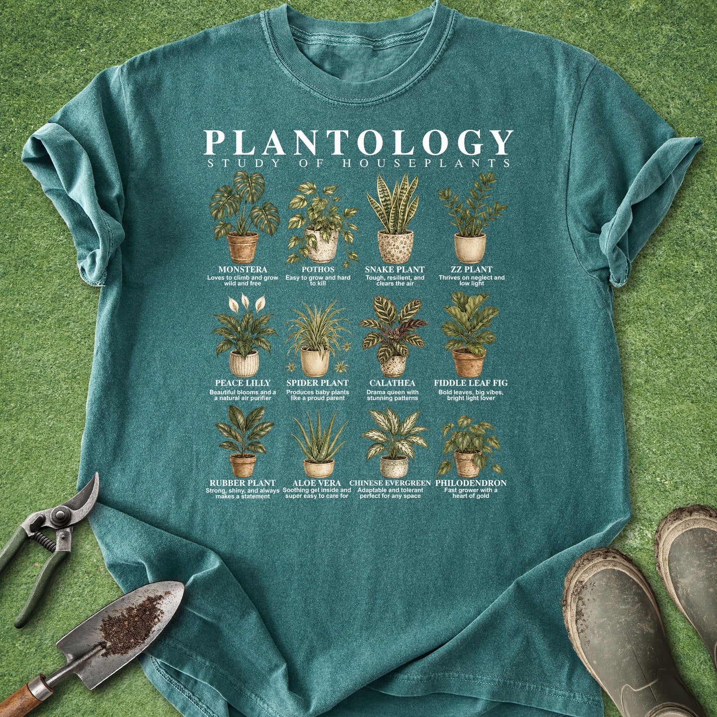 A teal short-sleeve graphic T-shirt laid flat on green grass with gardening tools and boots around it. The shirt features a large white and illustrated plant chart titled "PLANTOLOGY: STUDY OF HOUSEPLANTS" with multiple labeled houseplant drawings.