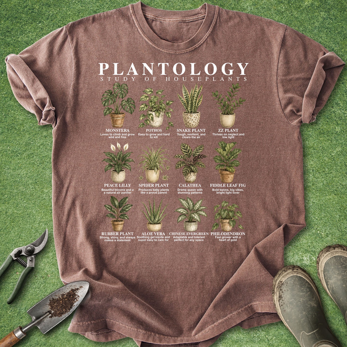 A brown short-sleeve graphic T-shirt laid flat on green grass-like background, with gardening tools and boots around it. The shirt reads 'PLANTOLOGY: STUDY OF HOUSEPLANTS' and shows a grid of illustrated houseplants with plant names beneath each one.