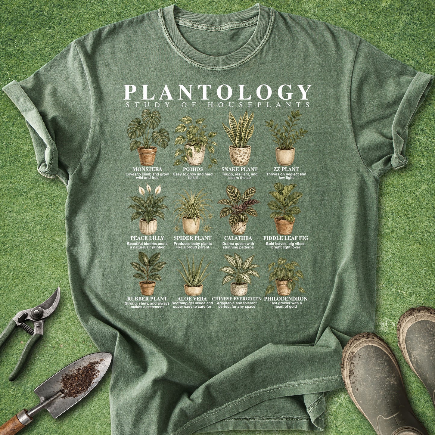 An olive green graphic t-shirt laid flat on a green grass-like surface, surrounded by gardening props including pruning shears, a trowel, and boots. The shirt features a white 'PLANTOLOGY STUDY OF HOUSEPLANTS' title and a grid of illustrated potted houseplants with plant names underneath.
