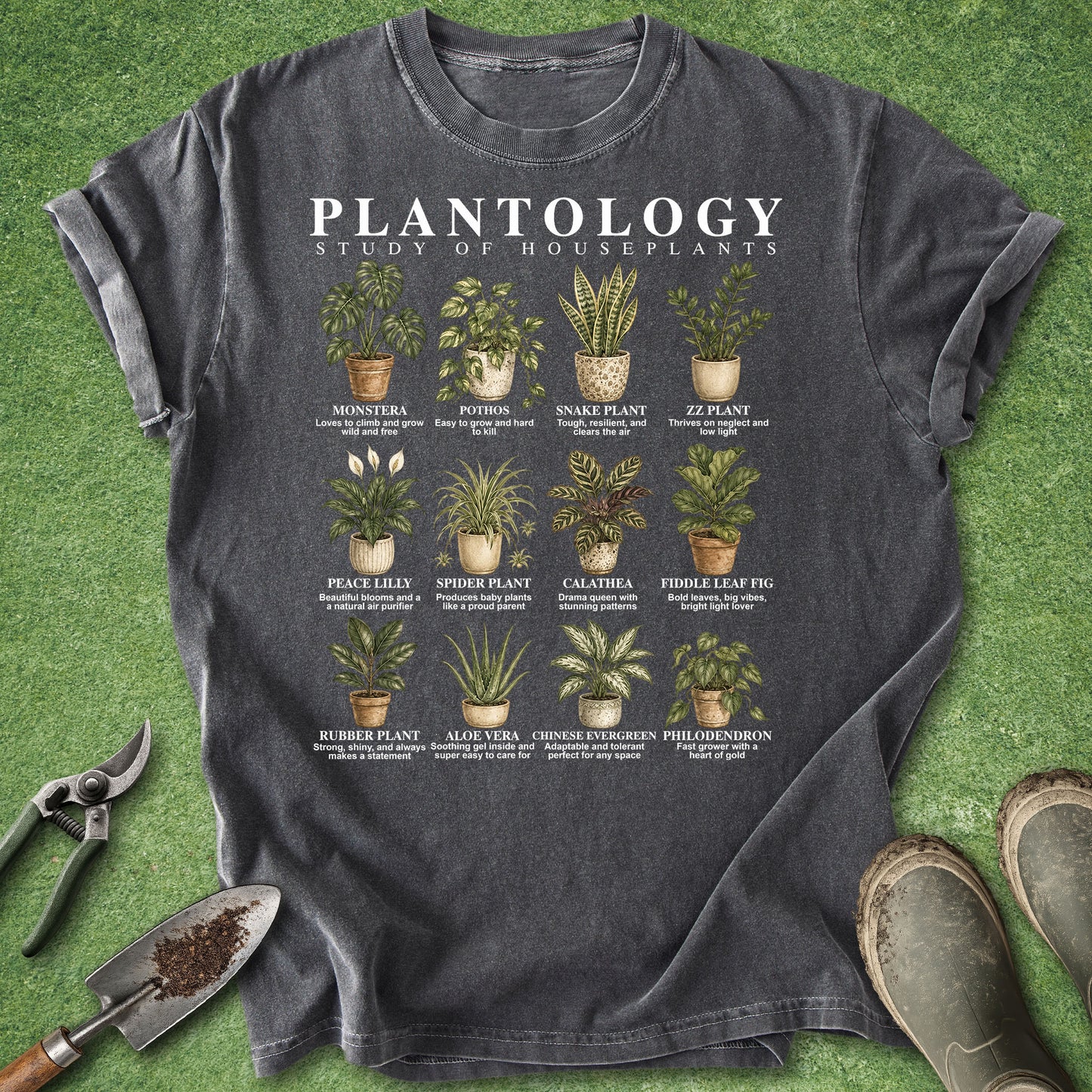 A dark charcoal gray graphic t-shirt laid flat on green grass-like artificial turf, surrounded by gardening props including pruning shears, a trowel, and boots. The shirt features a white 'PLANTOLOGY' title and a grid of illustrated houseplants with their names and care notes.