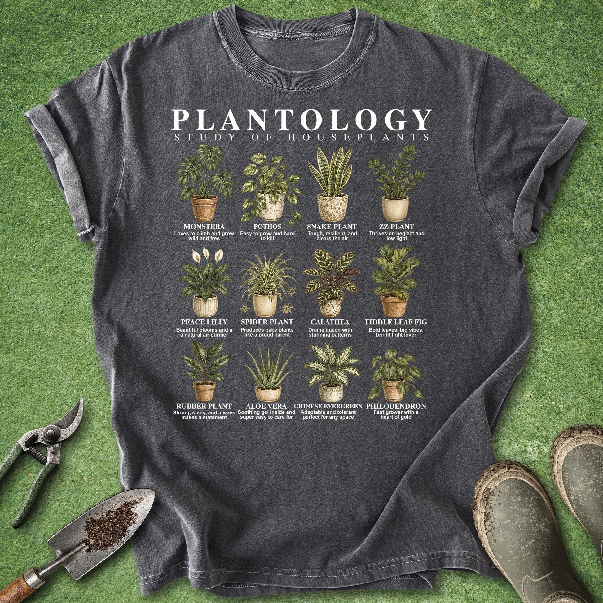 A dark charcoal gray graphic t-shirt laid flat on green grass-like artificial turf, surrounded by gardening props including pruning shears, a trowel, and boots. The shirt features a white 'PLANTOLOGY' title and a grid of illustrated houseplants with their names and care notes.